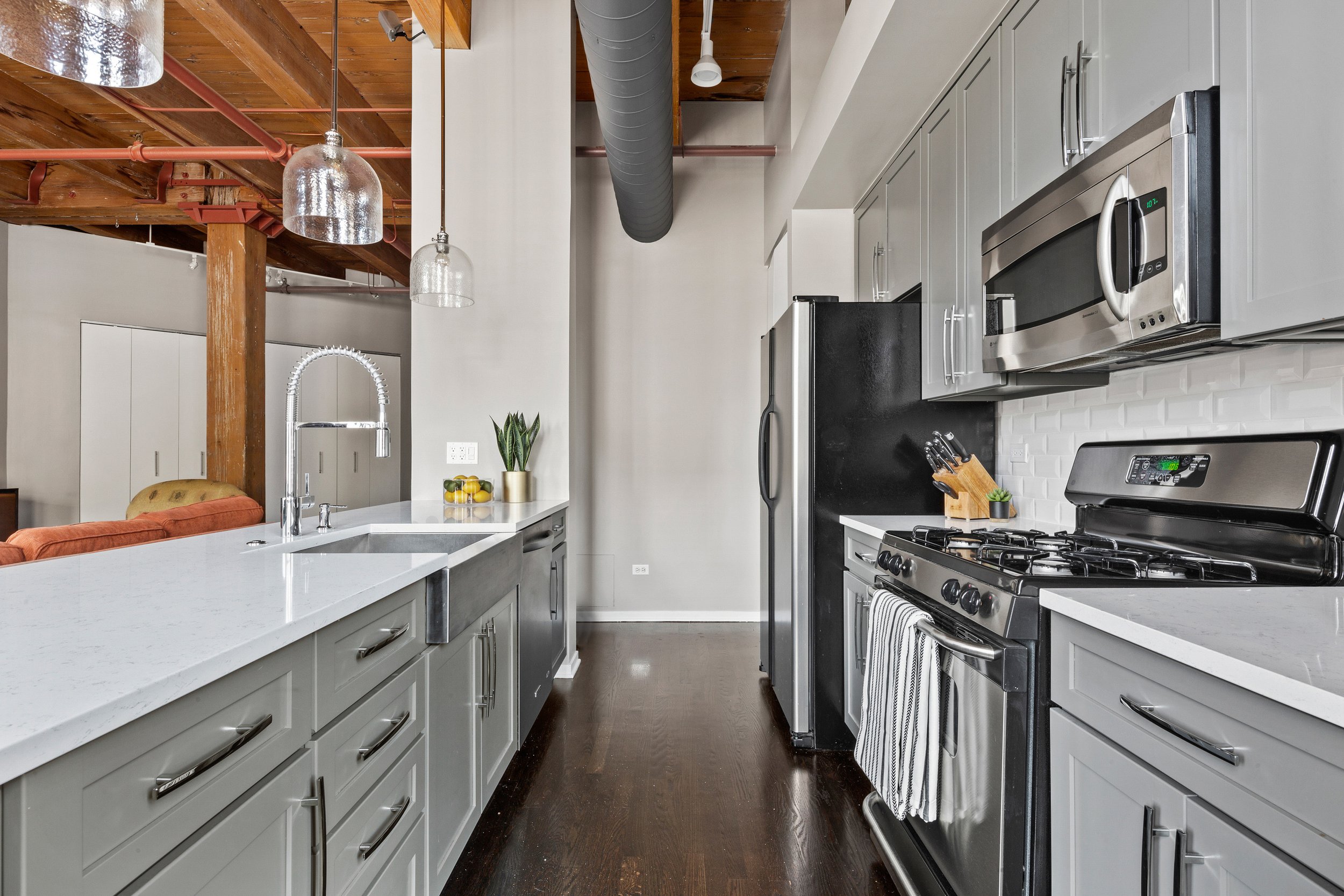Modern kitchen with white countertops, gray cabinets, stainless steel appliances including a microwave, oven, and refrigerator, dark wood floors, and exposed wooden beams and pipes on the ceiling.