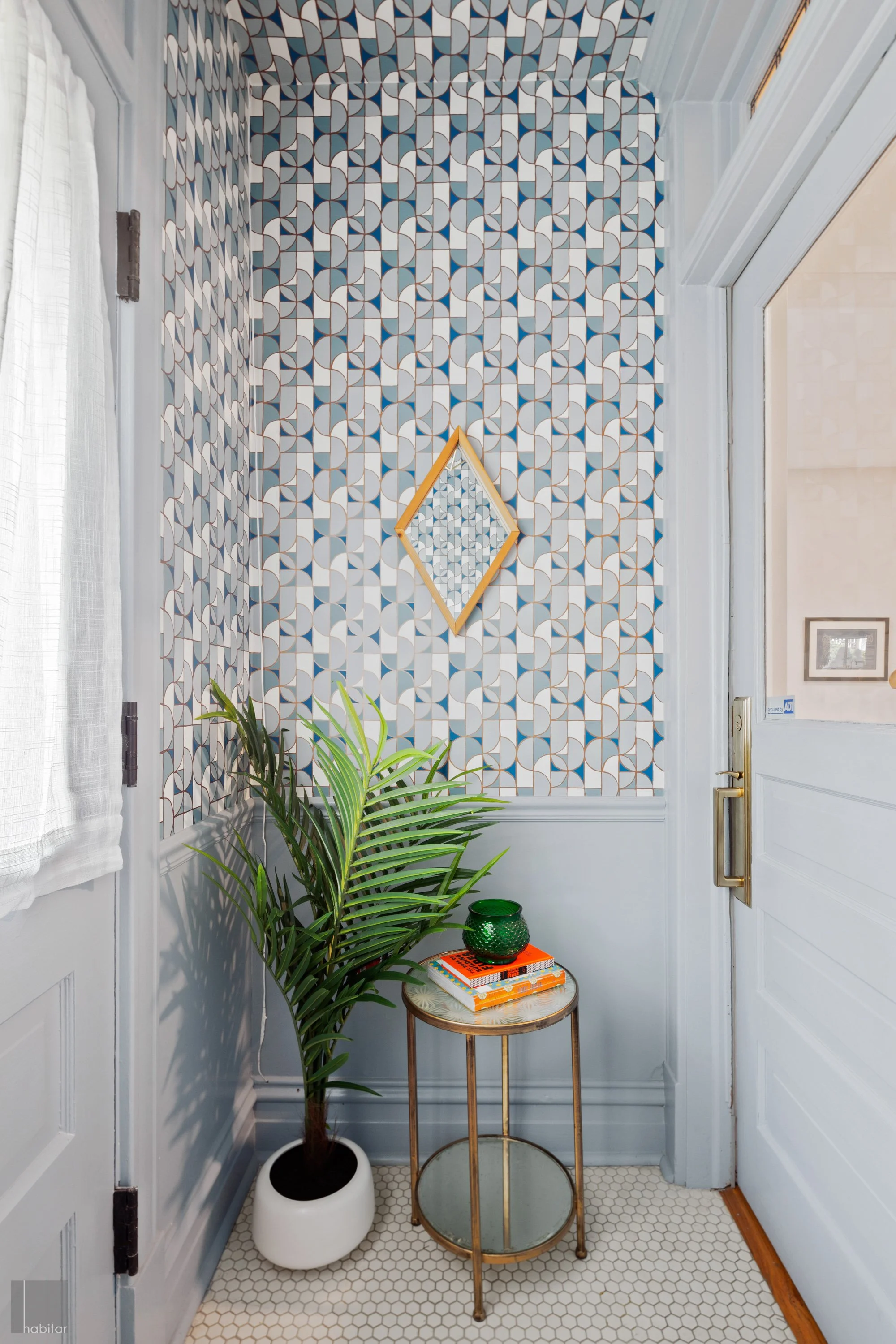 A cozy entryway with a patterned blue and white wallpaper, a white door with a brass handle, a small round table with a glass top and gold frame, decorated with a green glass bowl and stacked books, a large potted palm and a white curtain on the wind