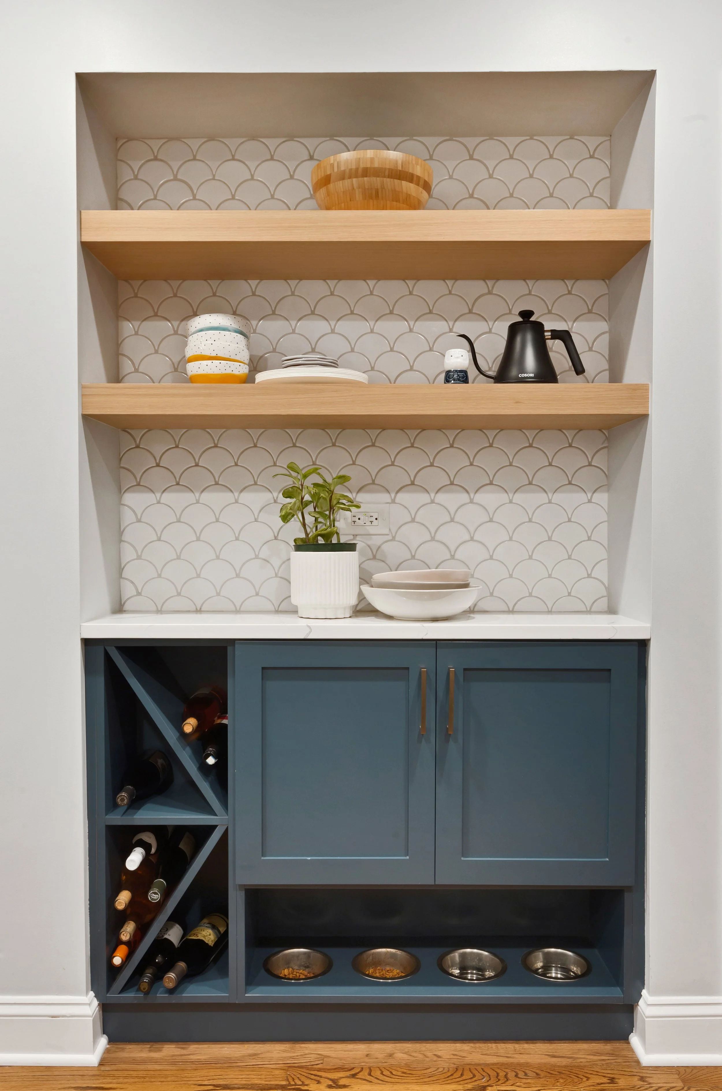 Built-in kitchen display with open shelving, a white countertop, and blue cabinetry, featuring a potted plant, bowls, plates, a black electric kettle, cups, and wine bottles stored in diagonal compartments.