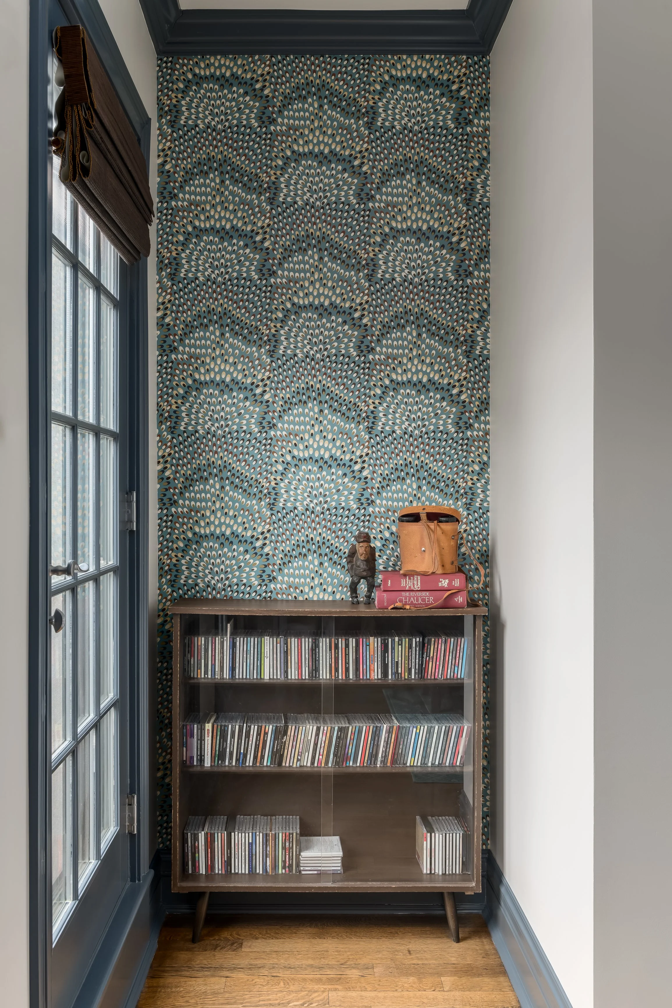Small wooden cabinet filled with CDs beneath a wall with blue, white, and black patterned wallpaper, topped with books and figurines, in a corner next to a window with blue trim.