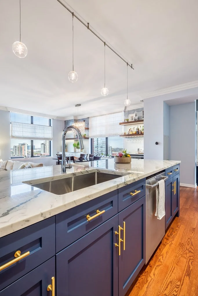 Modern kitchen with navy blue cabinets, white marble countertops, and gold hardware. There is a stainless steel sink with a curved faucet, and a bowl of fruit on the counter. The space is brightly lit with three hanging pendant lights and large windo