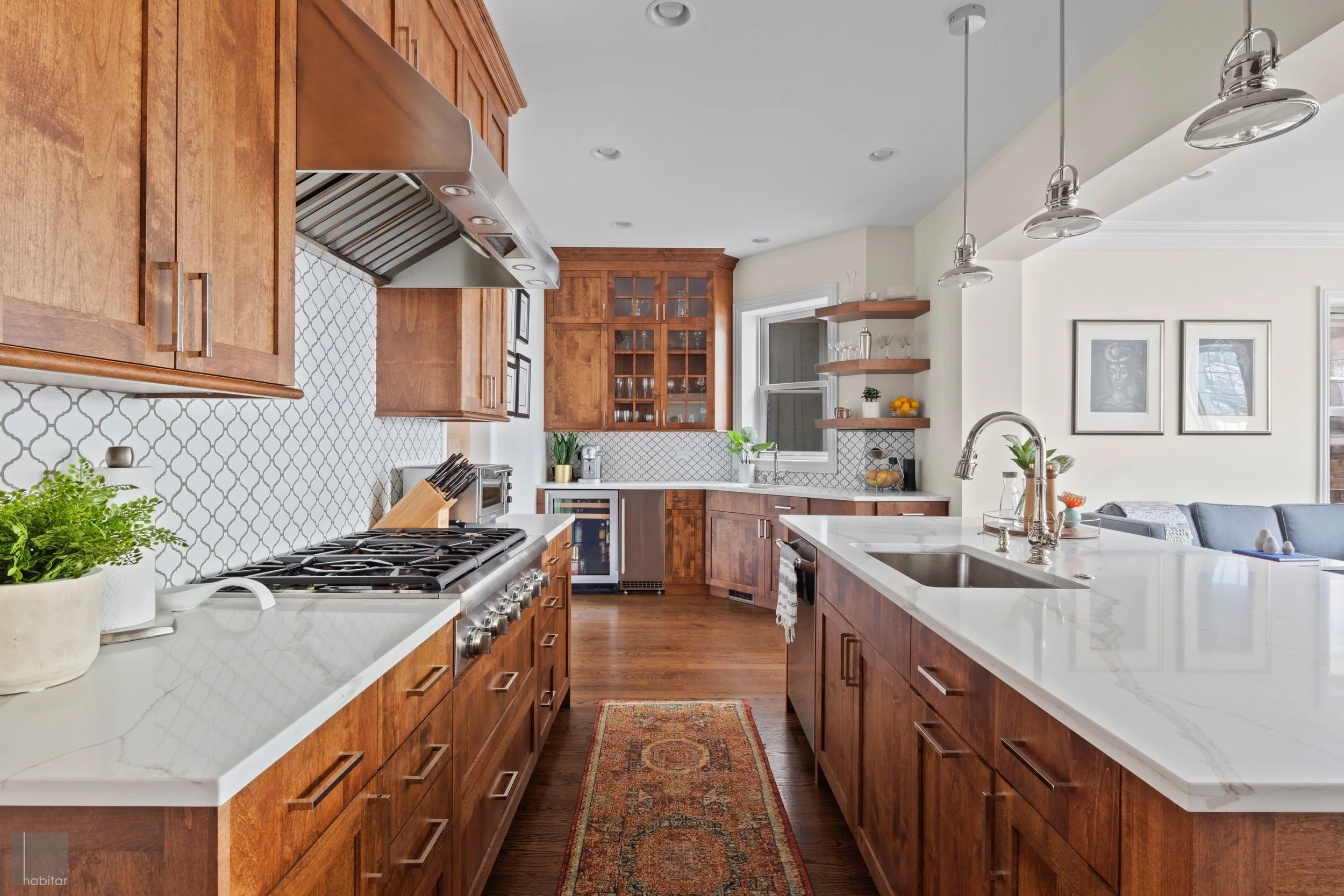 Modern kitchen with wooden cabinets, white marble countertops, and a backsplash with a white geometric tile pattern. The space includes a stove, sink, and small decorative plants, with open shelves and a large window letting in natural light. A livin