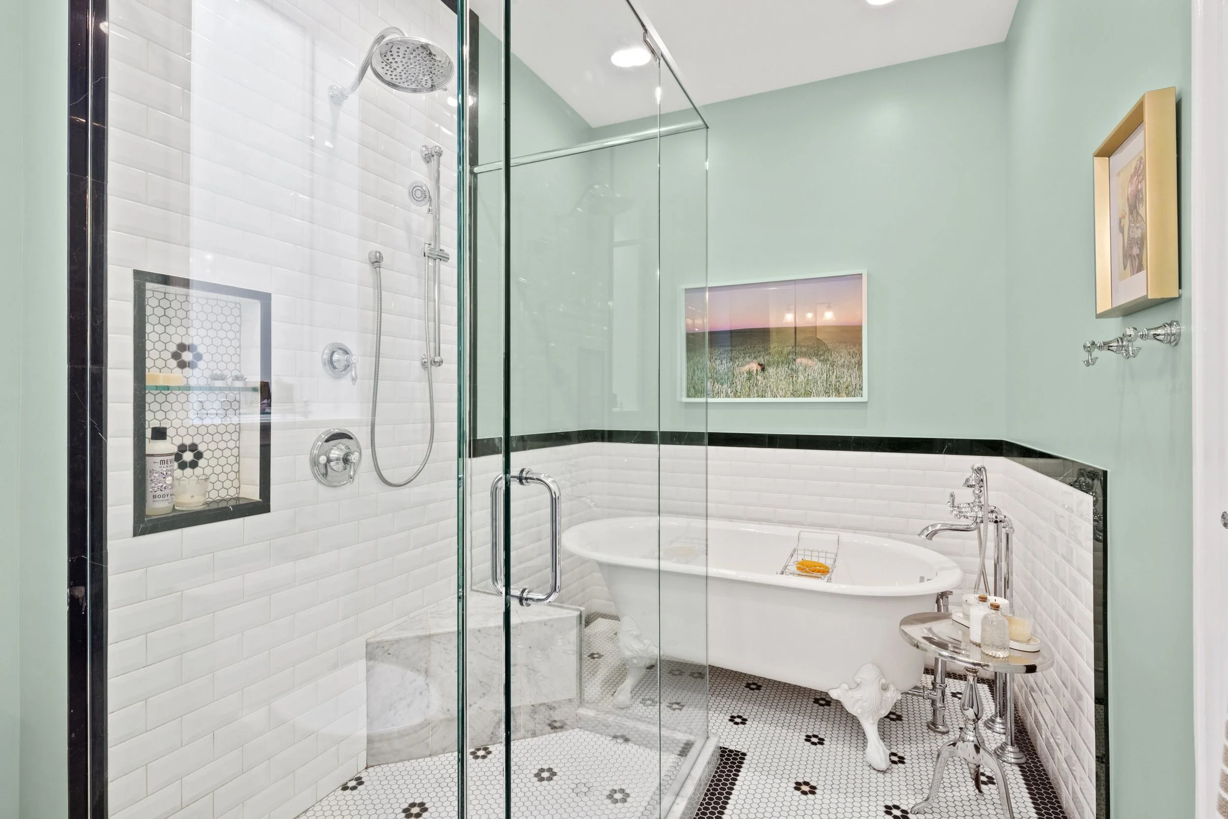 Bathroom with glass shower enclosure, vintage clawfoot bathtub, black and white tile floor, mint green walls, framed landscape photo, and artwork.