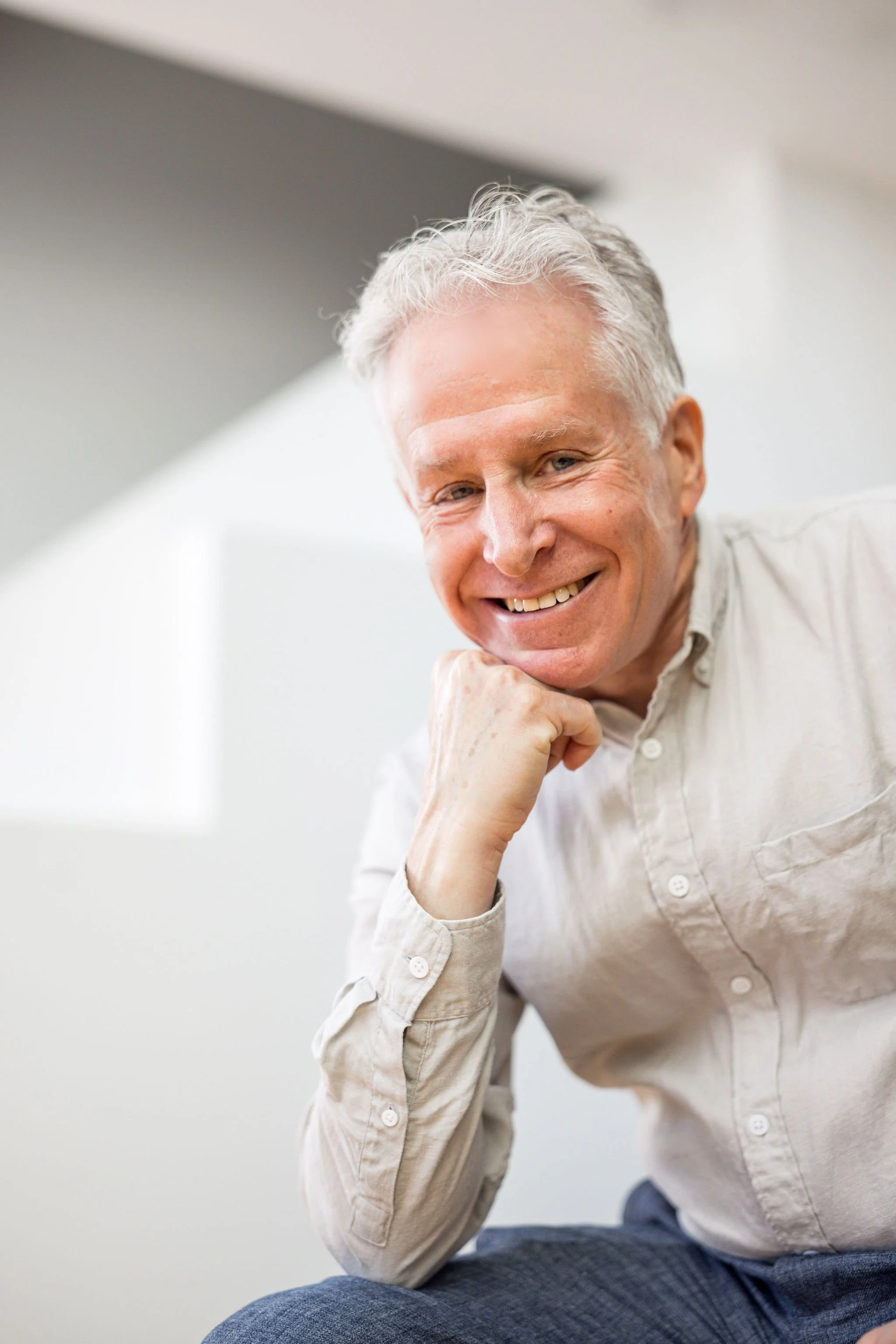 Smiling older man with gray hair, wearing a beige shirt, sitting and resting his chin on his hand indoors.