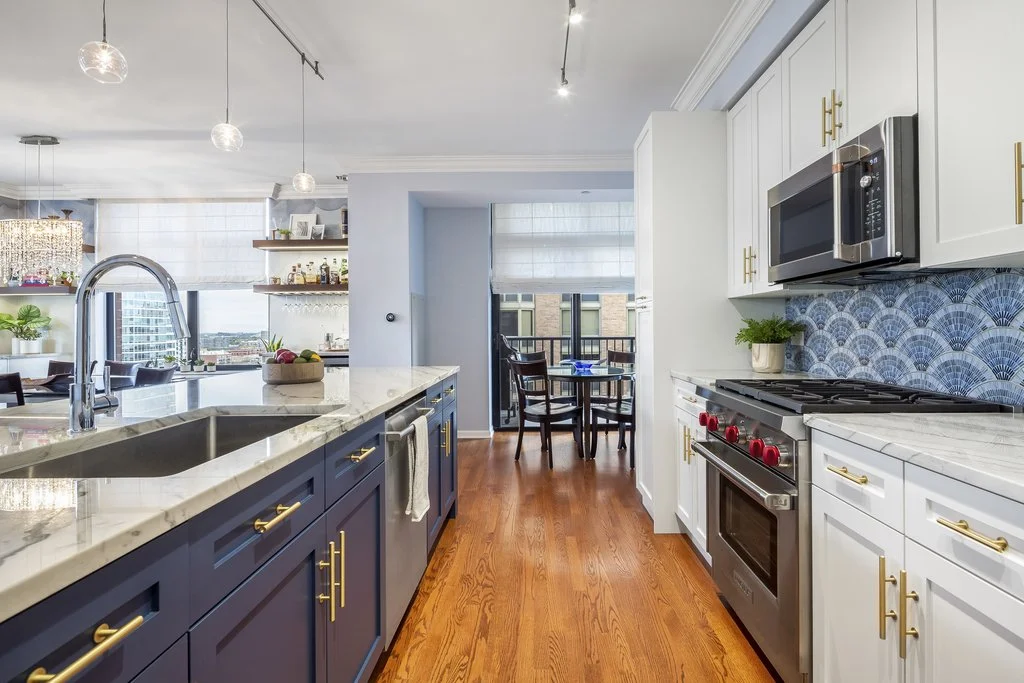 Modern kitchen with white cabinets, stainless steel appliances, blue backsplash, wooden floors, and a small dining area with a window view.