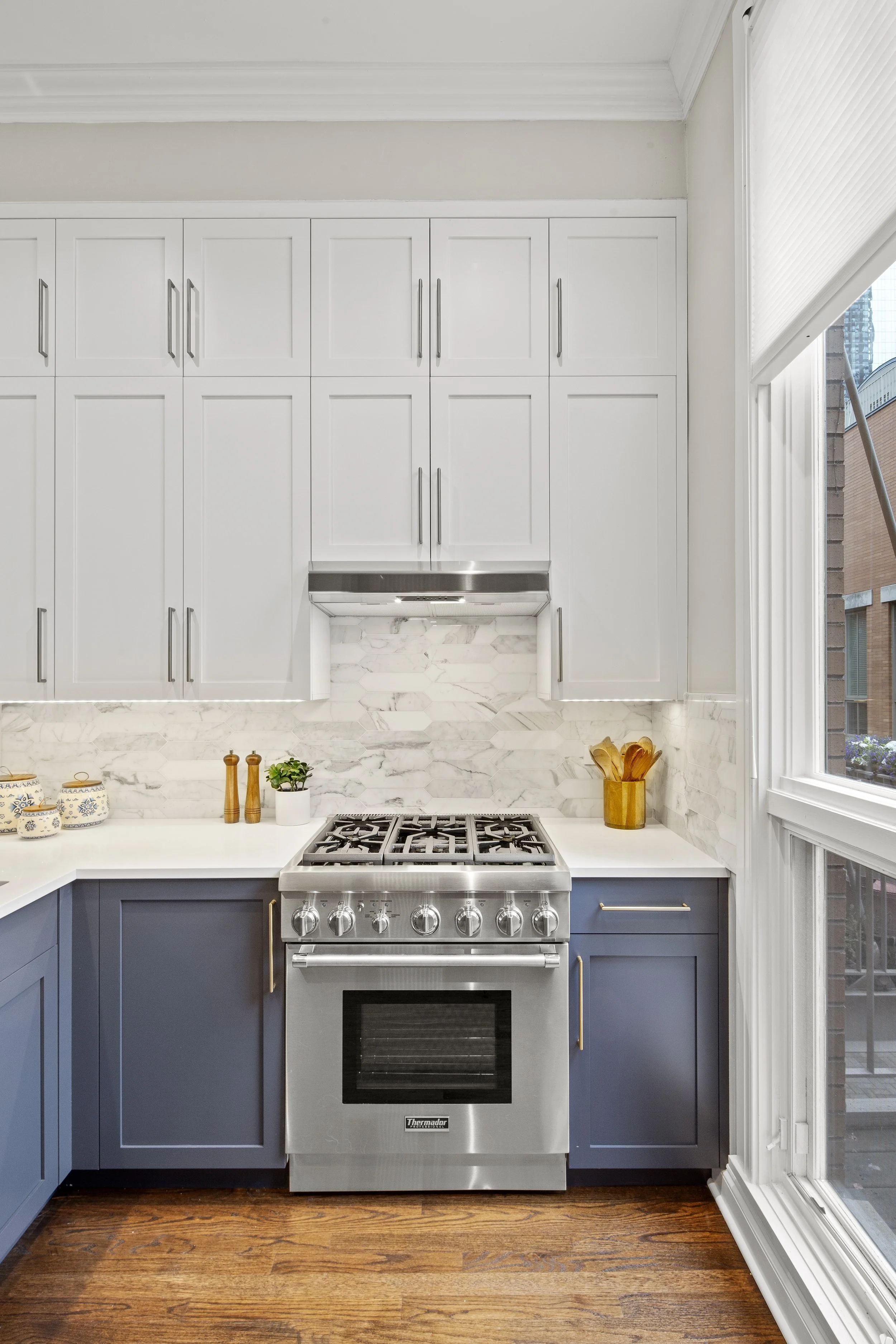Modern kitchen with white upper cabinets, dark blue lower cabinets, marble backsplash, and stainless steel stove near a window.