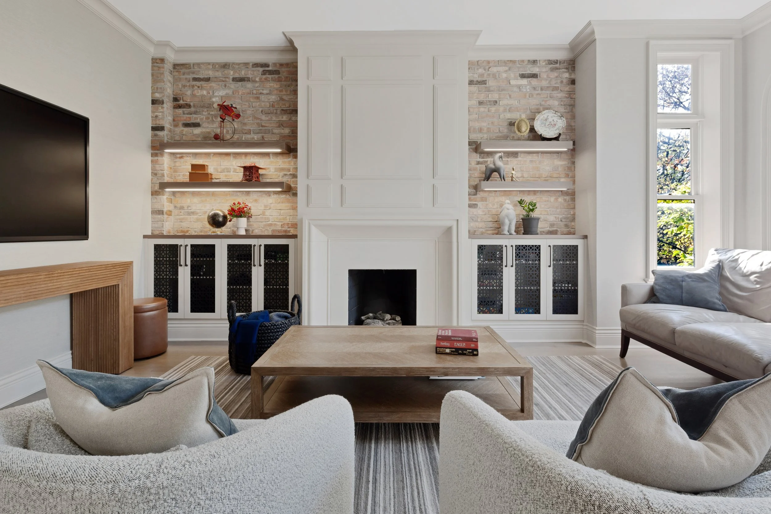 Living room with a white fireplace, brick wall, built-in shelves with decorations, large window, white sofa with pillows, wooden coffee table with books, and striped rug.
