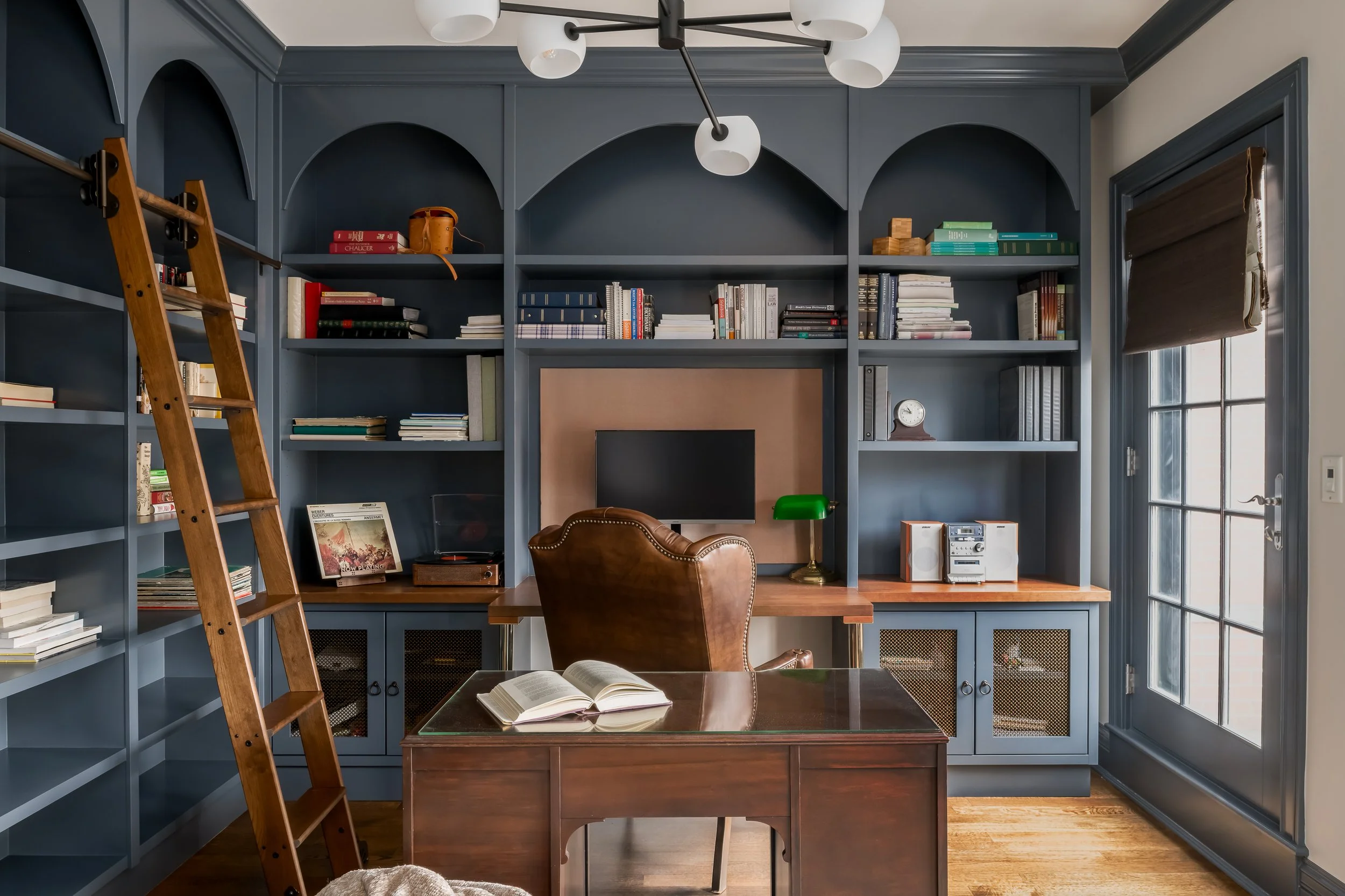 Home office with blue built-in shelves, a wooden desk, a leather chair, a ladder, and a window with a brown roller blind.