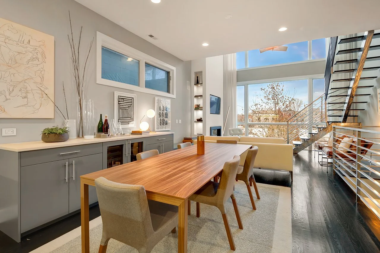 Contemporary living and dining area with light grey walls, a large wooden dining table with six chairs, a sideboard with decorative bottles and plants, abstract wall art, and large windows letting in natural light.