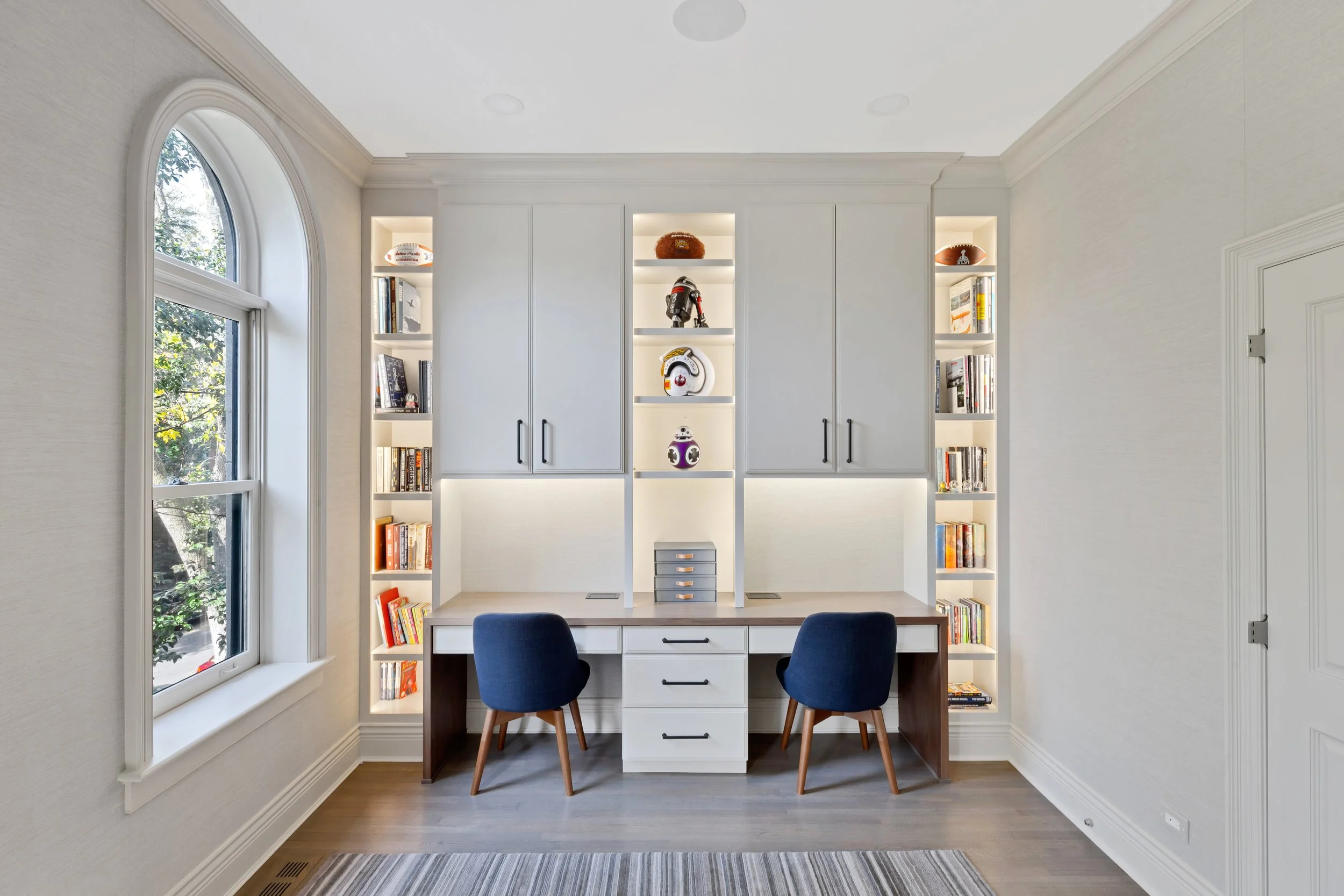 Home office with a built-in white desk, two navy chairs, built-in shelving with books and Star Wars themed collectibles, a large window with arched top, and a striped rug on wood floor.