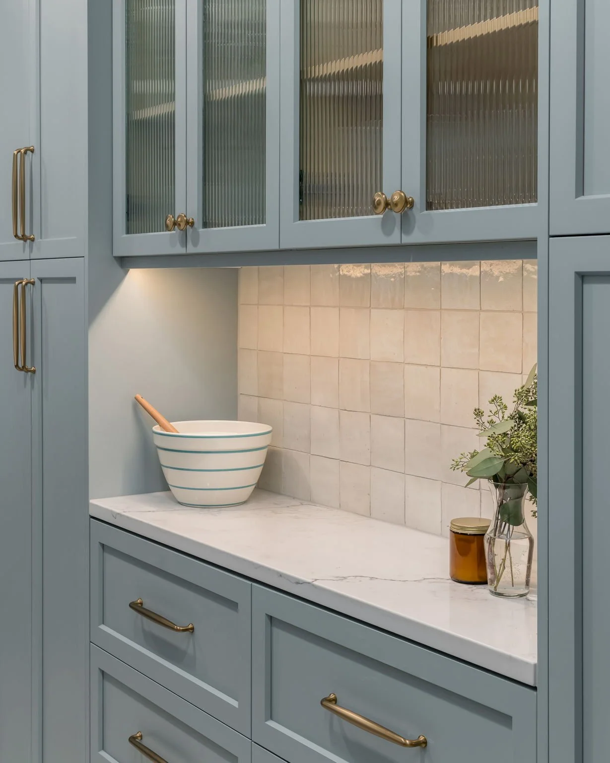 Blue kitchen cabinets with brass handles, a white marble countertop, and a beige tiled backsplash. There is a striped ceramic bowl with a wooden spoon, a small orange jar, and a glass vase with greenery.
