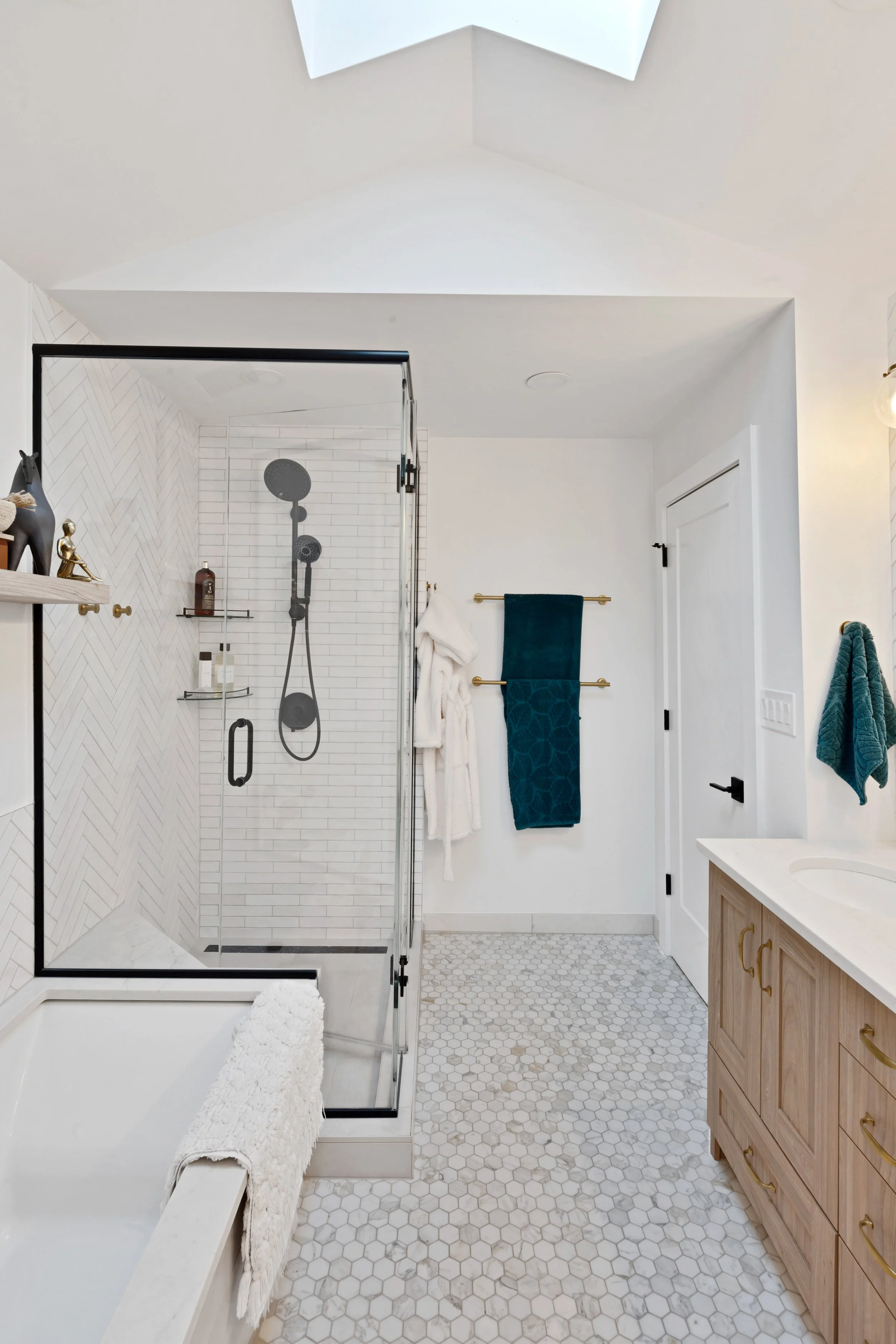 Modern bathroom with walk-in shower, white hexagon tile flooring, wooden vanity with brass hardware, and white walls with teal towels.