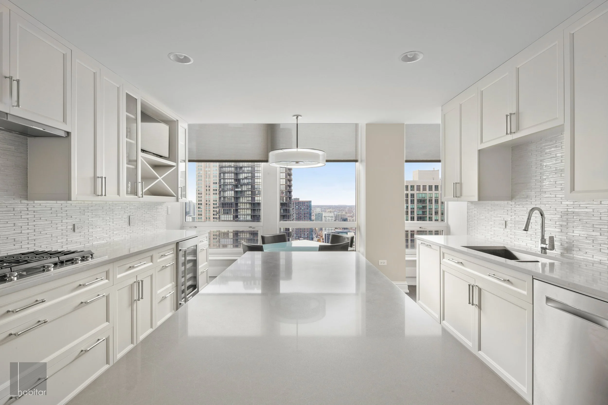 Modern white kitchen with large island, white cabinets, brick backsplash, and city view from windows.