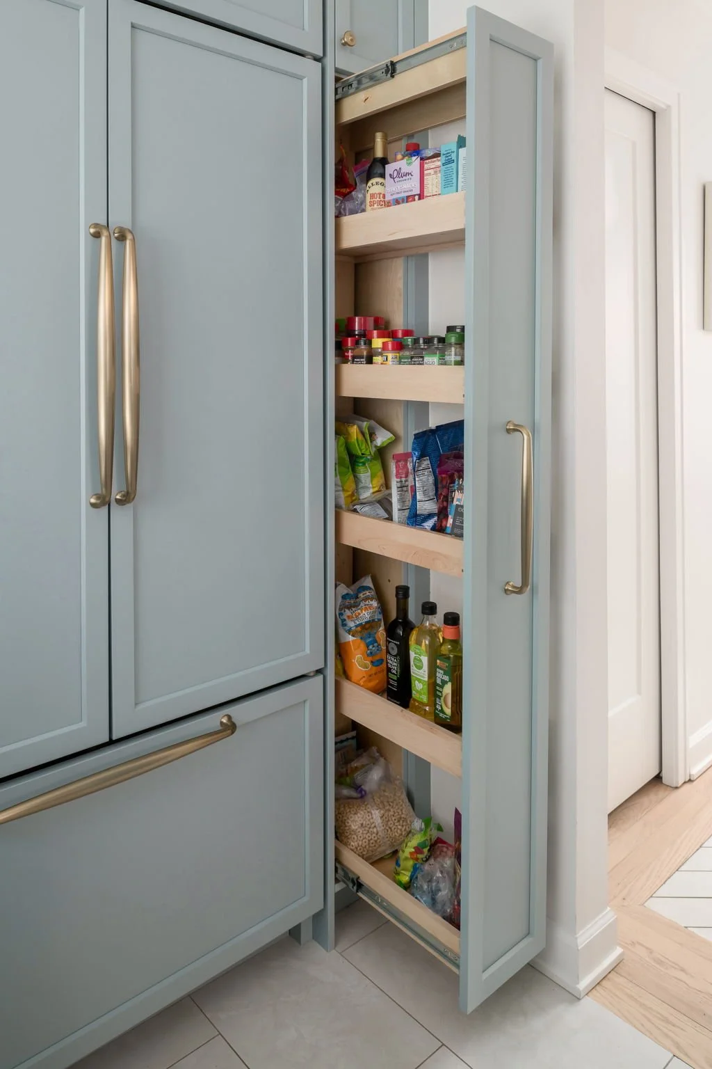 Open pull-out pantry with snacks, spices, and condiments in a kitchen with light blue cabinets.