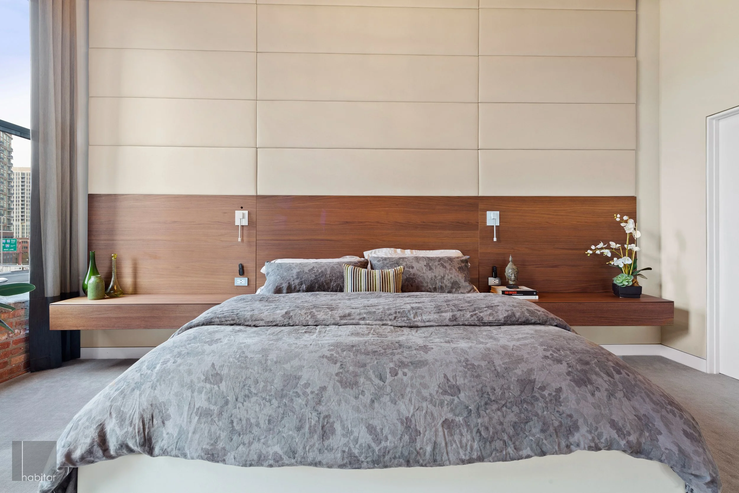 Modern bedroom with a large bed, gray bedding, and decorative pillows. Wooden floating nightstands on each side of the bed hold vases and decorative objects. The wall behind the bed features beige cushioned panels, with wall-mounted reading lights.