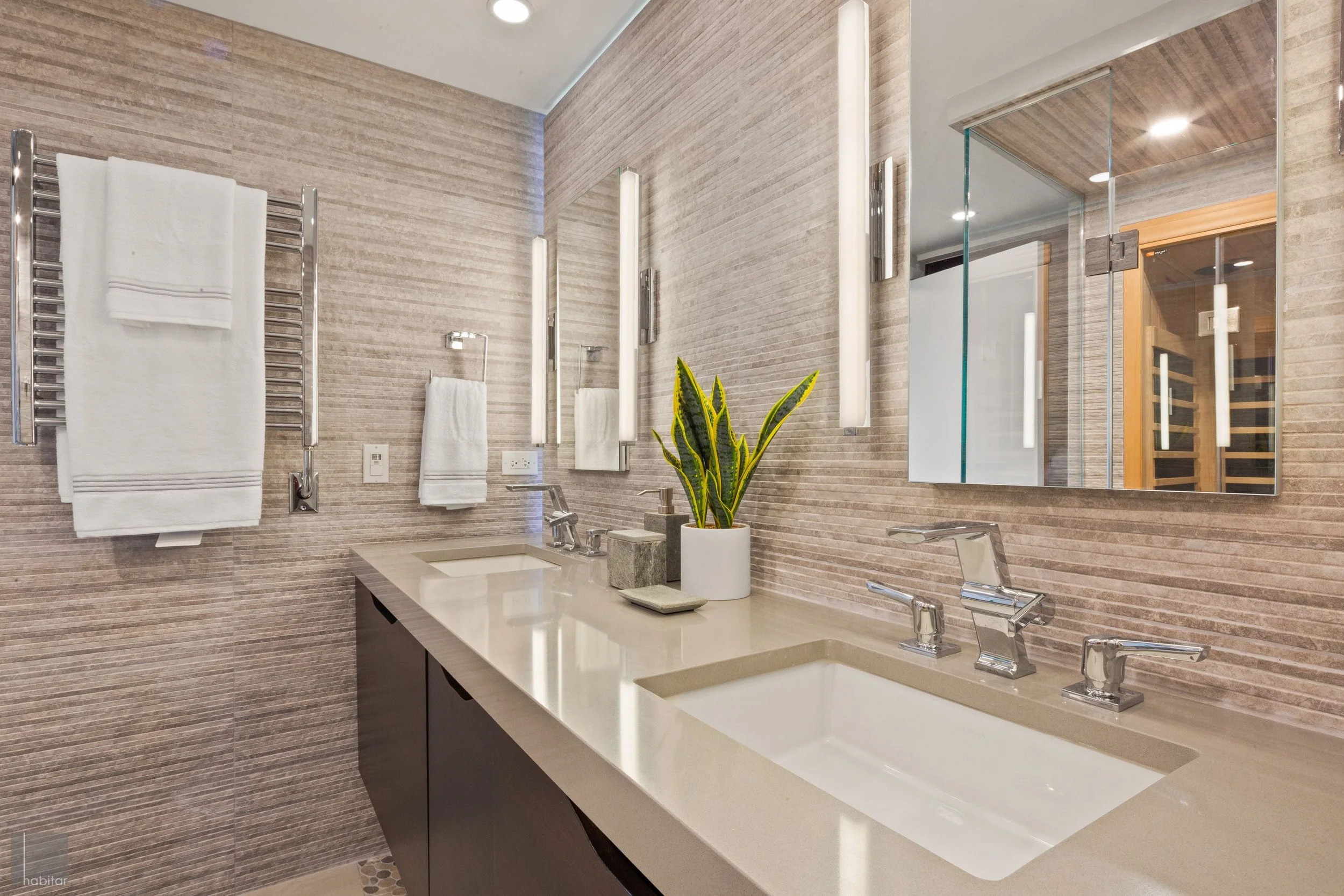 Modern bathroom with dual sinks, large mirrors, wall-mounted lighting, towels on a heated towel rail, and a potted plant on the countertop.