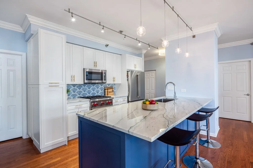 Modern kitchen with white cabinets, a blue island, marble countertop, stainless steel appliances, pendant lights, and hardwood floors.