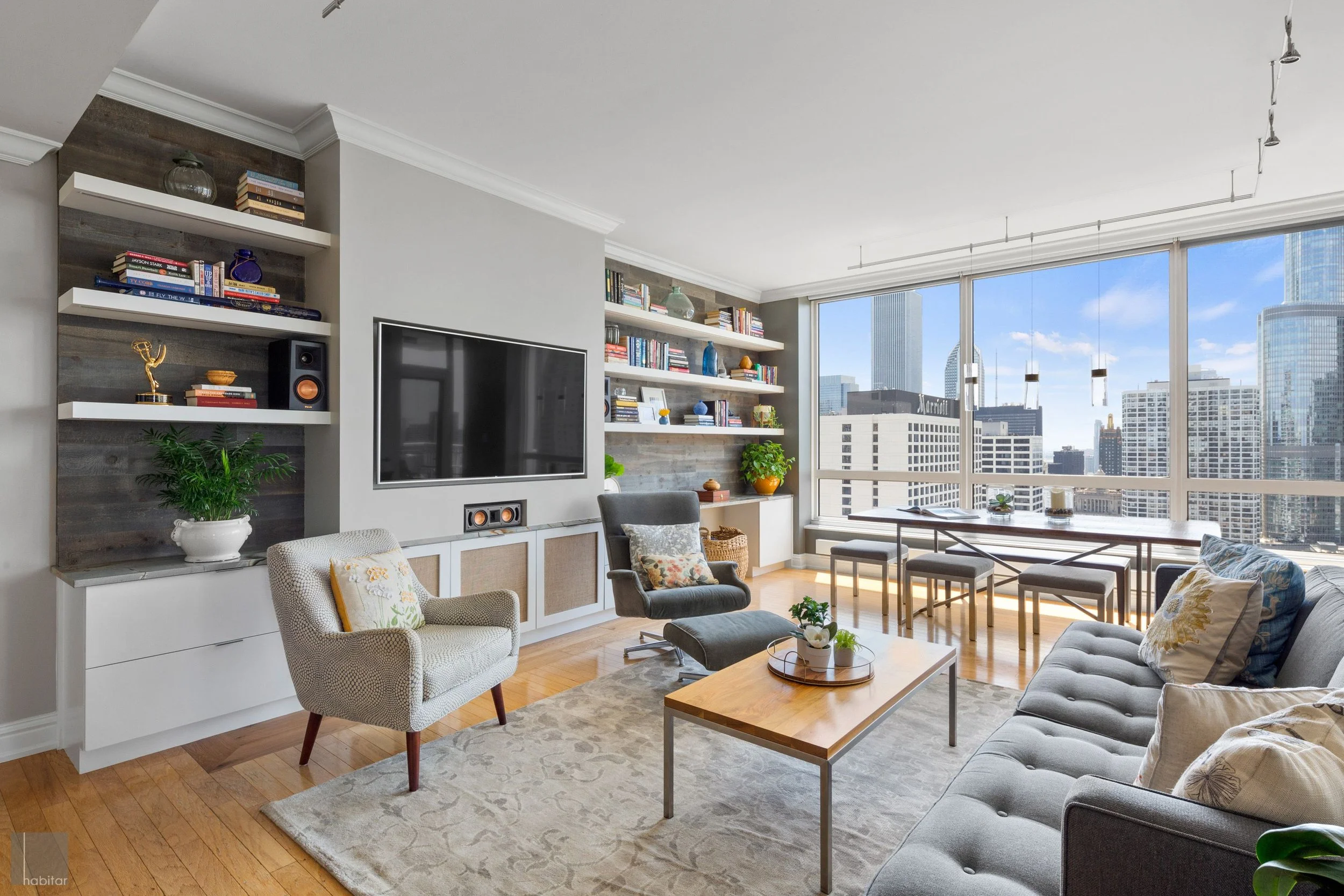 Modern living room with large windows overlooking city skyline, featuring a gray sofa with decorative pillows, a white armchair, a wooden coffee table, a gray accent chair with an ottoman, built-in shelves with books and decor, and a large flat-scree