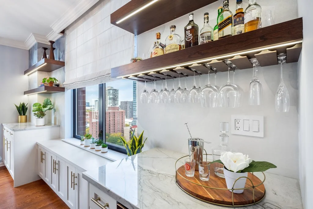 Kitchen bar area with white marble countertop, hanging wine glasses, bottles of alcohol on a wooden shelf, potted plants, and city view through window.
