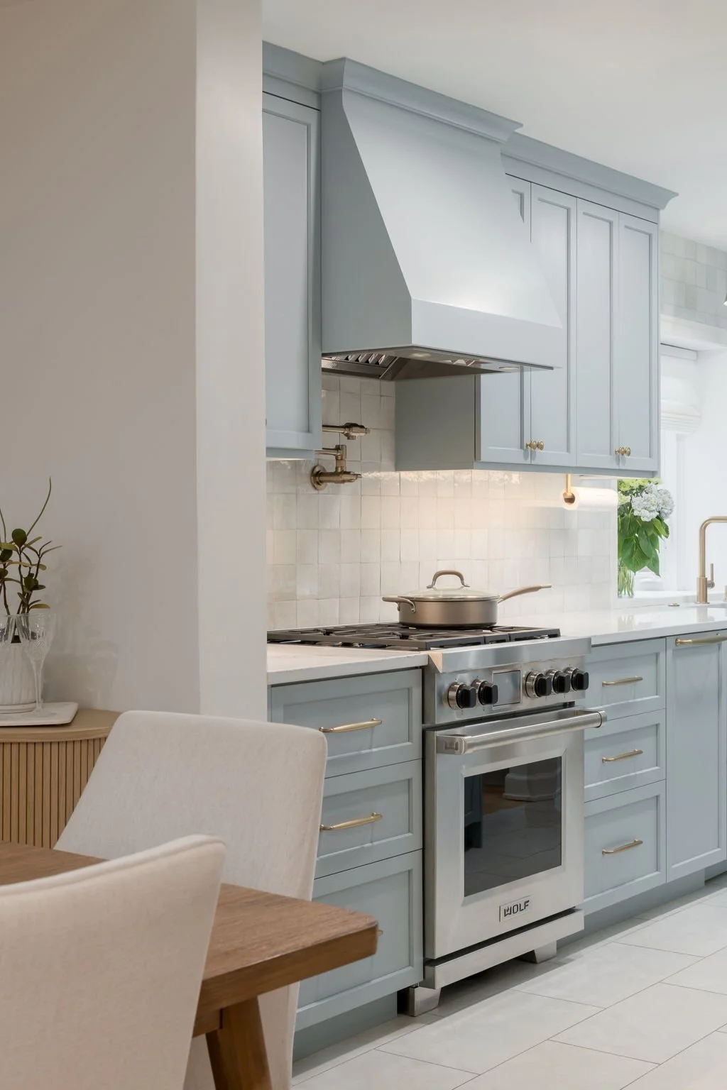 Light blue kitchen with stainless steel stove and range hood, white tiled backsplash, and white countertops. A pot is on the stove, and a vase with white flowers is near the window.