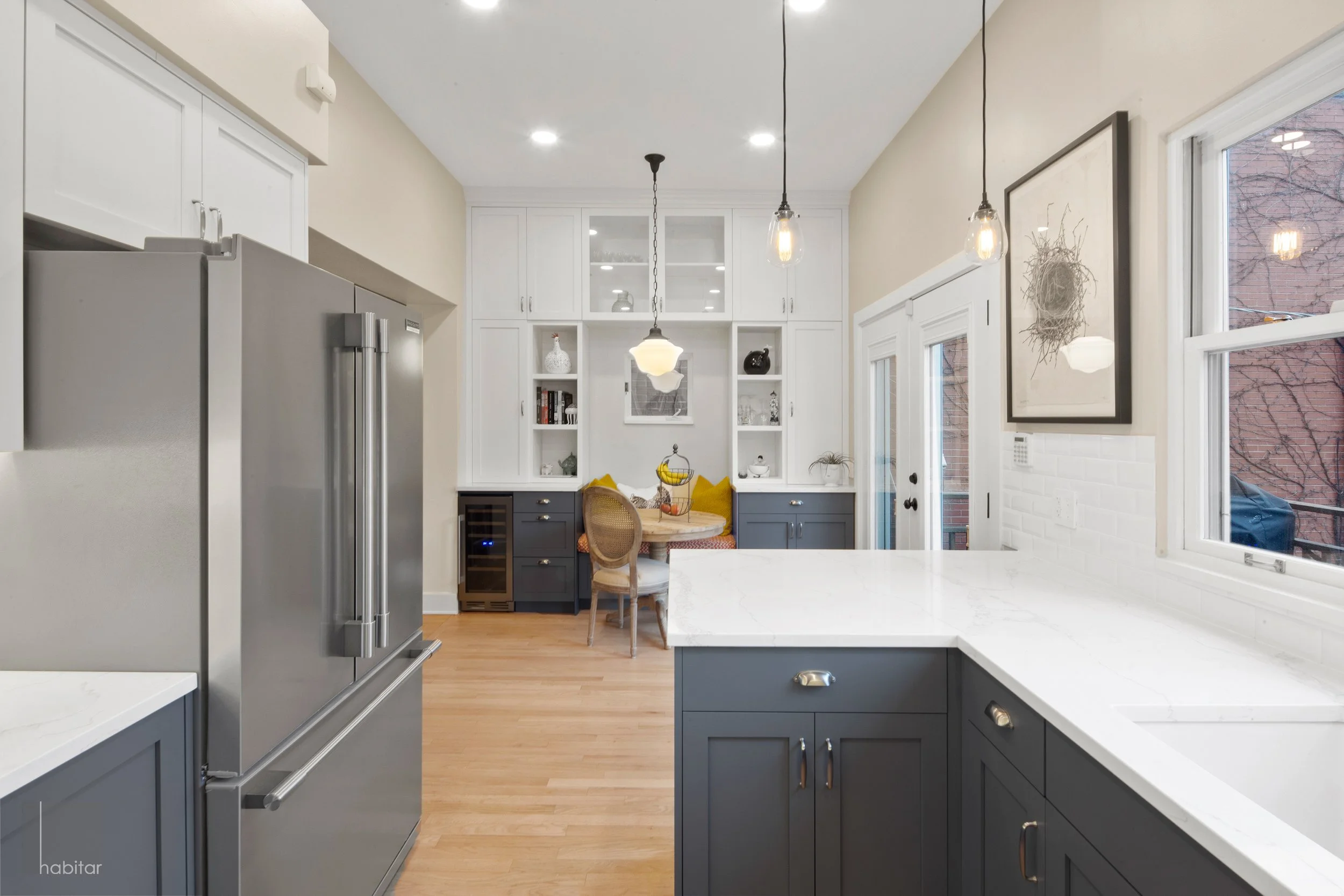 Modern kitchen with stainless steel refrigerator, white cabinets, gray lower cabinets, white marble countertops, and a breakfast nook with yellow pillows and a circular table near a built-in white cabinet with open shelves.