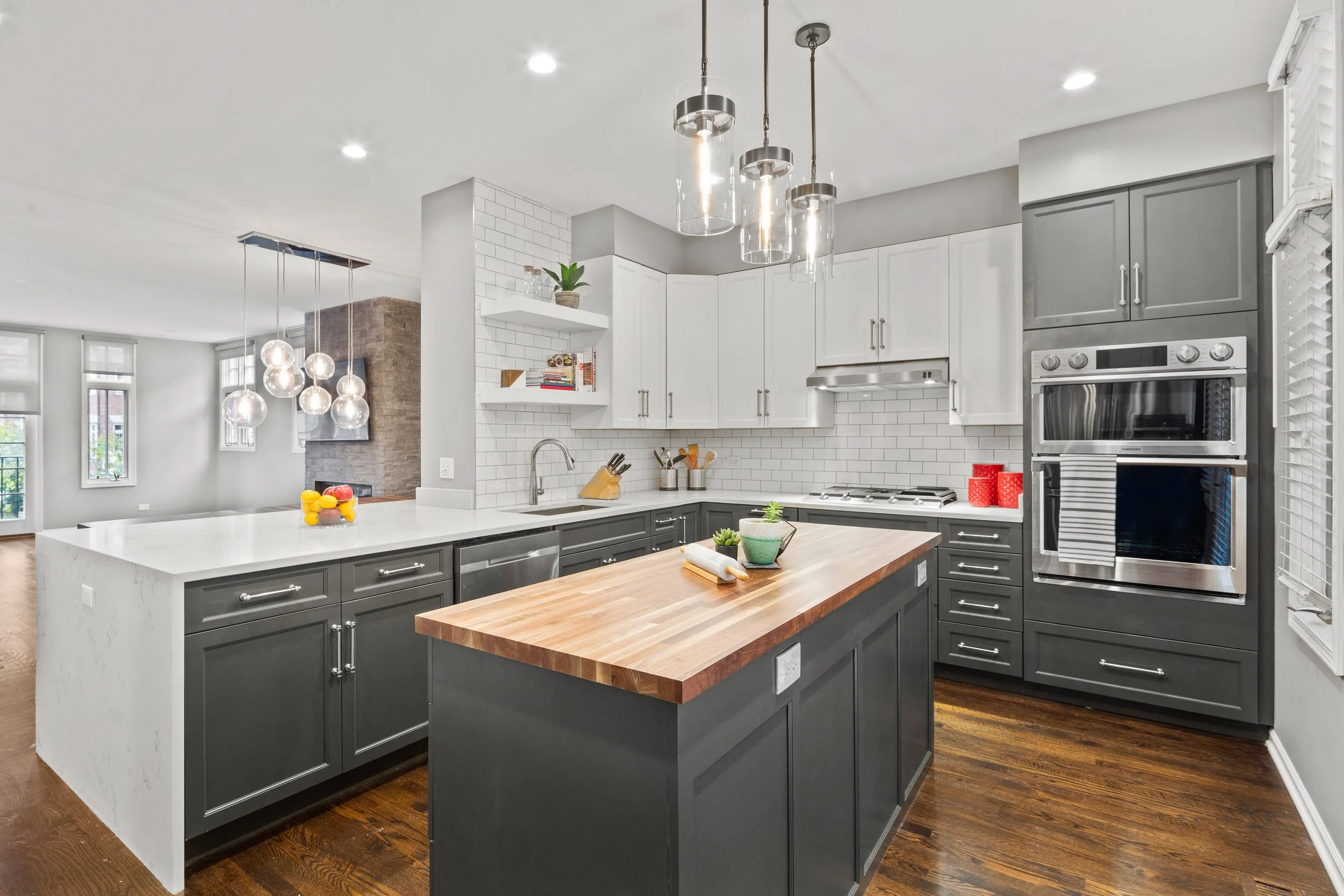 Modern kitchen with white upper cabinets, dark lower cabinets, a wooden island countertop, white subway tile backsplash, stainless steel appliances, and pendant lighting fixtures.