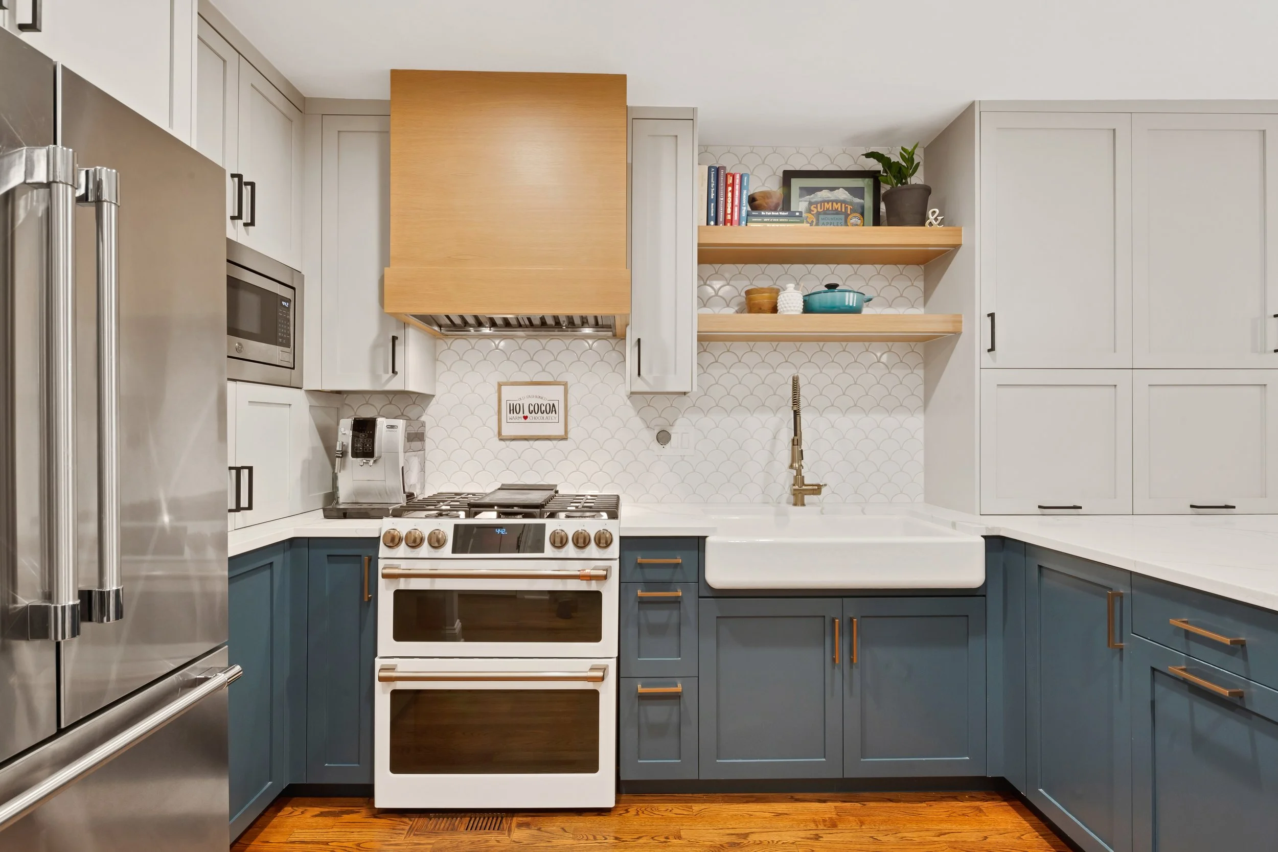 Modern kitchen with blue lower cabinets, white upper cabinets, white farmhouse sink, brass faucet, stainless steel refrigerator, stove, microwave, shelves with books and plants, hardwood floor, tiled backsplash.