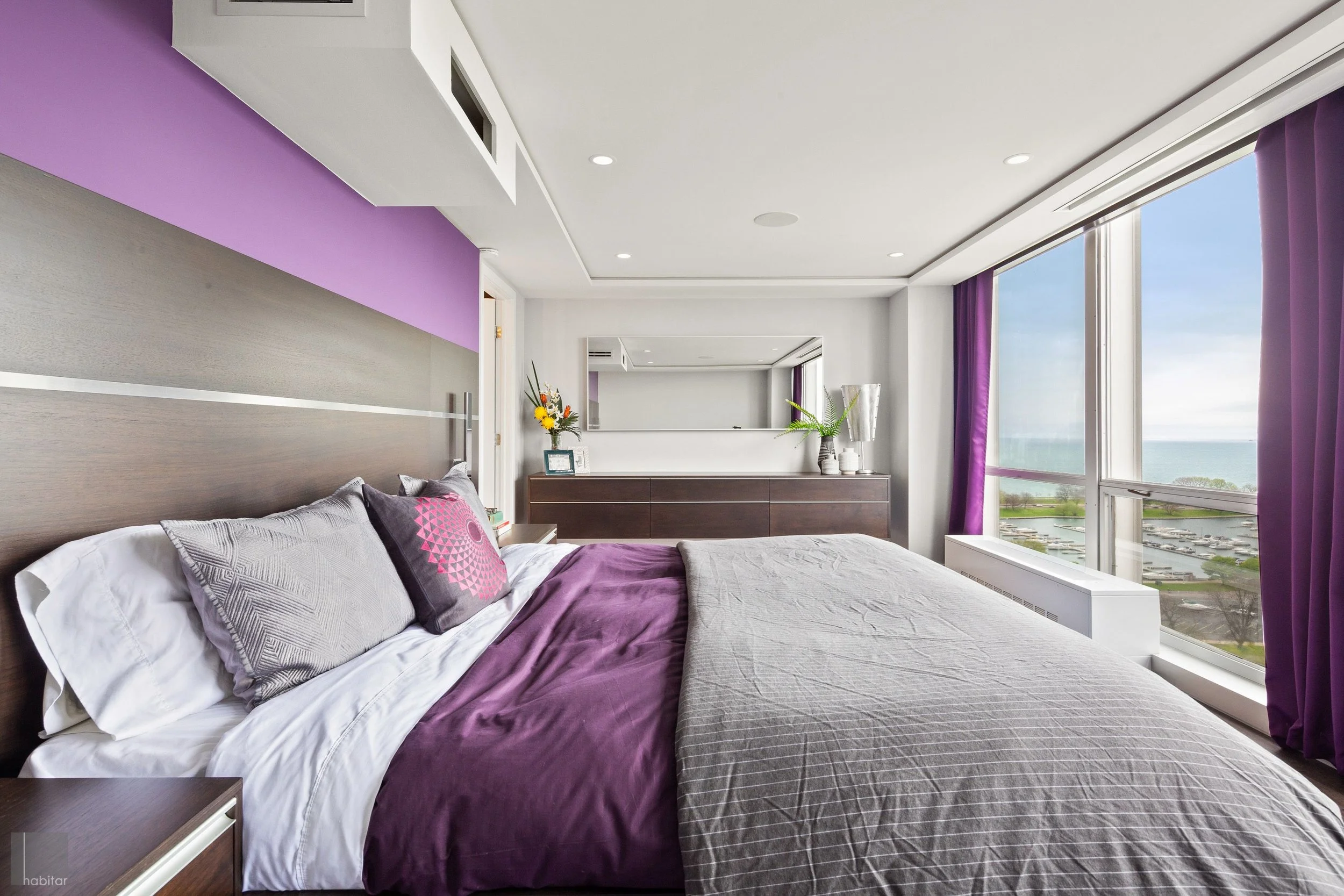 A modern hotel room with a large bed, purple and gray bedding, and pillows. There is a window with a view of a parking lot and water. A dresser with decorative items and a mirror is in the background.