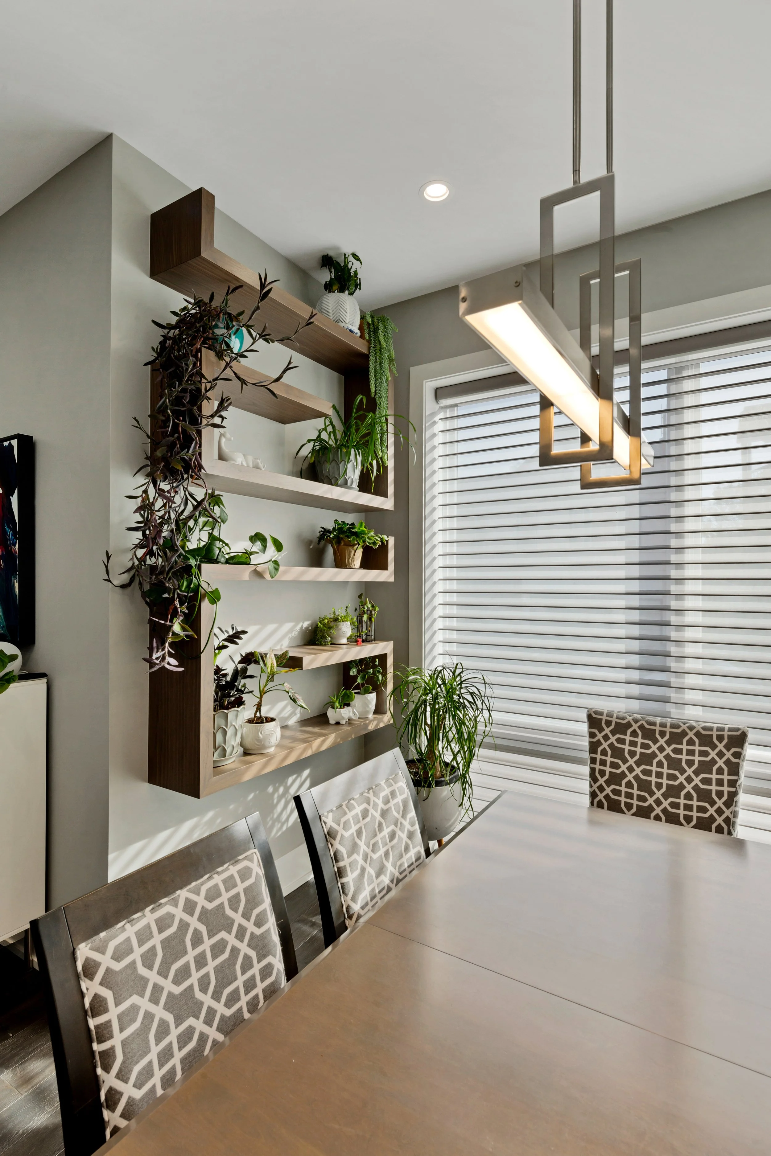 A modern dining room with a wooden table, patterned upholstered chairs, a hanging geometric light fixture, a wall-mounted wooden shelf with plants, and large windows with blinds.