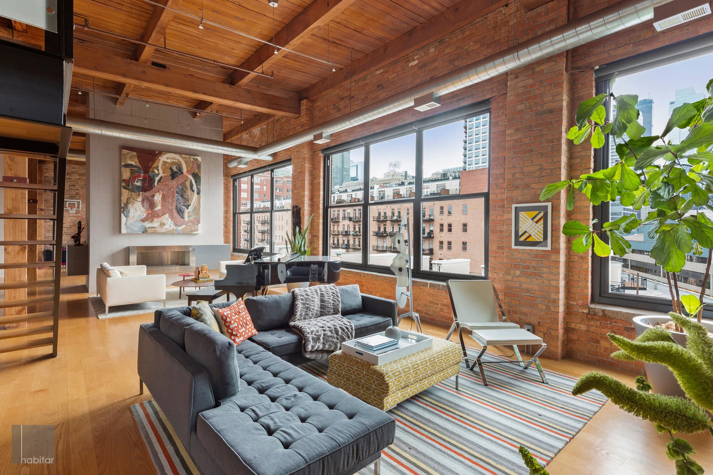 Modern loft apartment living room with exposed brick walls, large windows, wooden ceiling beams, and colorful furniture including a dark gray sectional sofa, striped area rug, yellow ottoman, and white lounge chair, with artwork on the walls and city buildings visible outside.