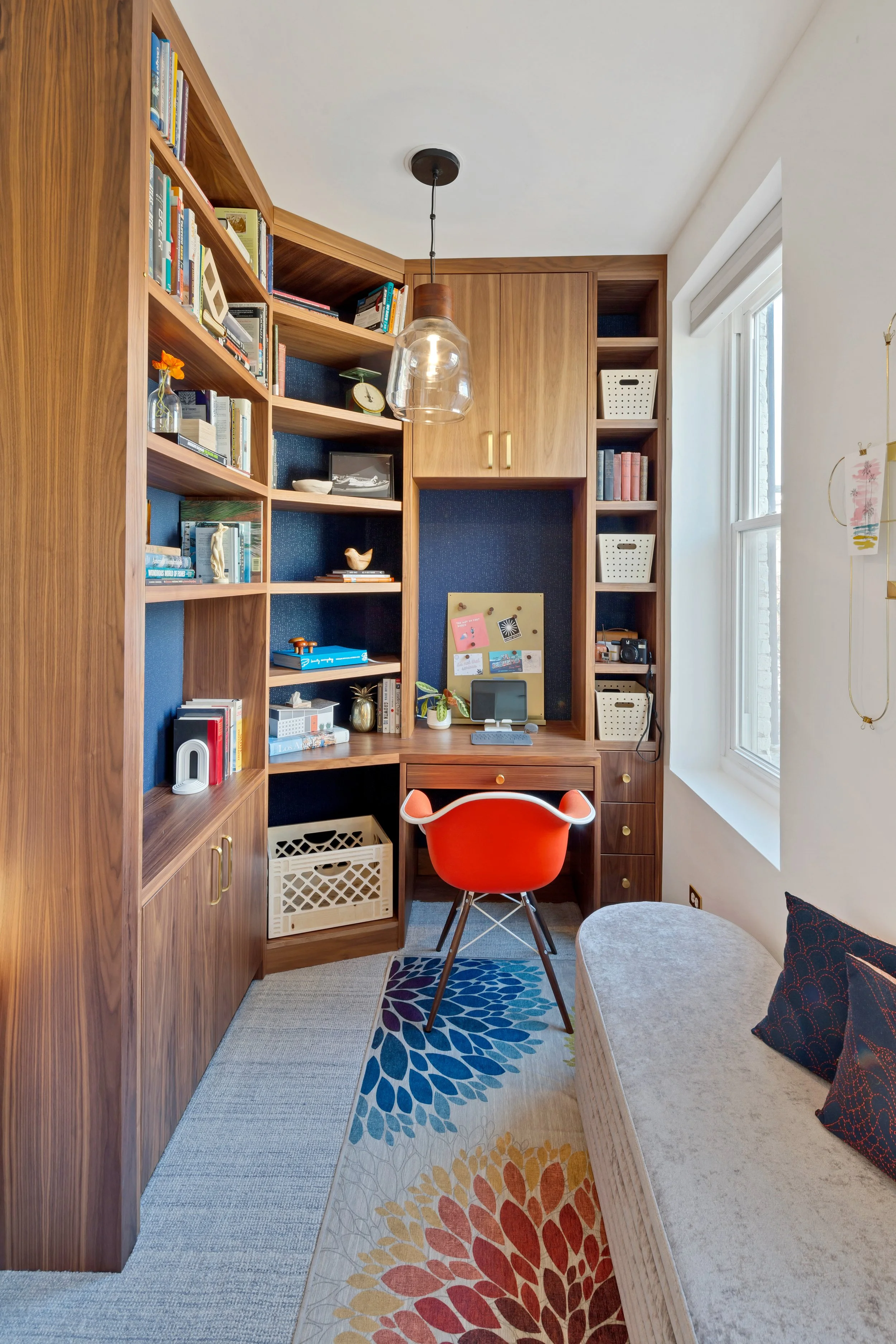 Home office with built-in wooden shelves, a red chair at a wooden desk, a window with white trim, and a patterned rug.
