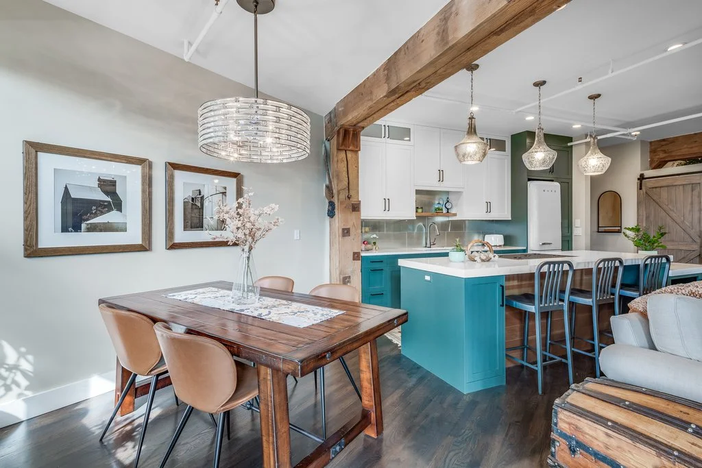 Open concept kitchen and dining area with white and teal cabinets, a wooden beam, three hanging pendant lights, a wooden dining table with tan chairs, and wall art.
