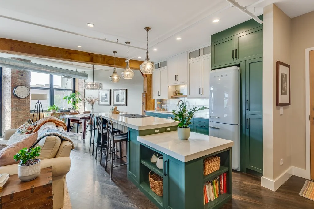 Open-concept living space with kitchen and dining area, featuring green and white kitchen cabinets, a white island with a potted plant, pendant lights, a dining table near large windows, and a cozy beige sofa with blankets and pillows.