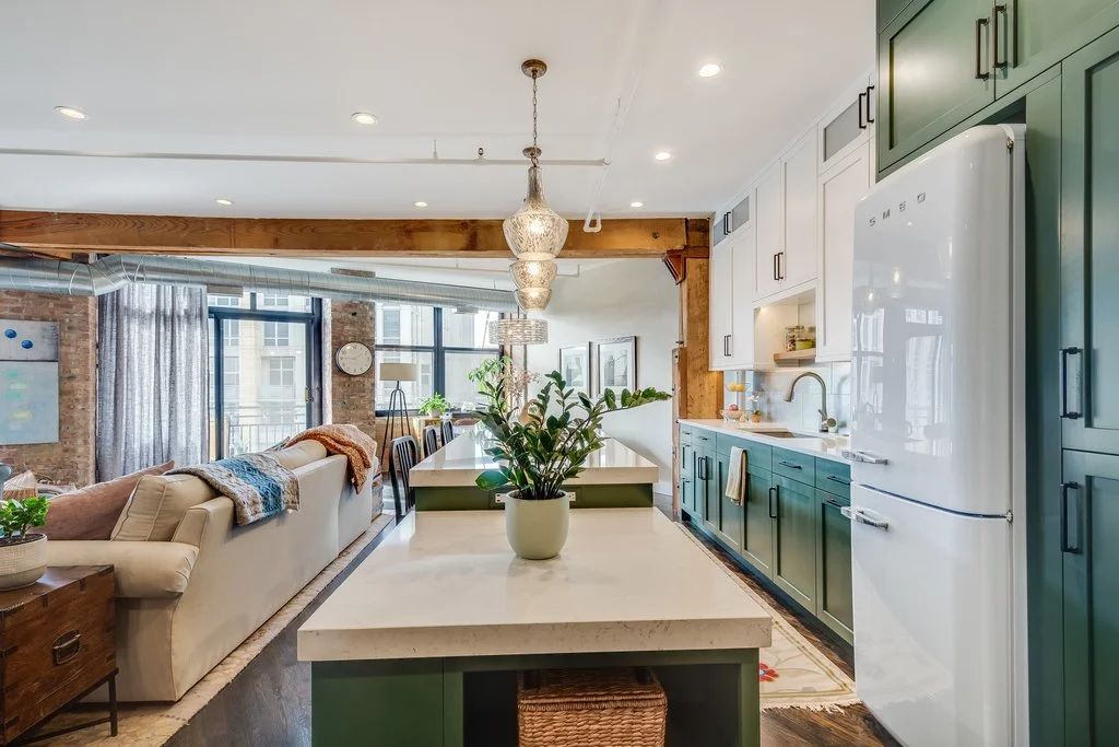 Open-concept living space with kitchen and living room, featuring white and green cabinets, a white refrigerator, a kitchen island with a plant, large windows, exposed brick wall, and cozy decor.