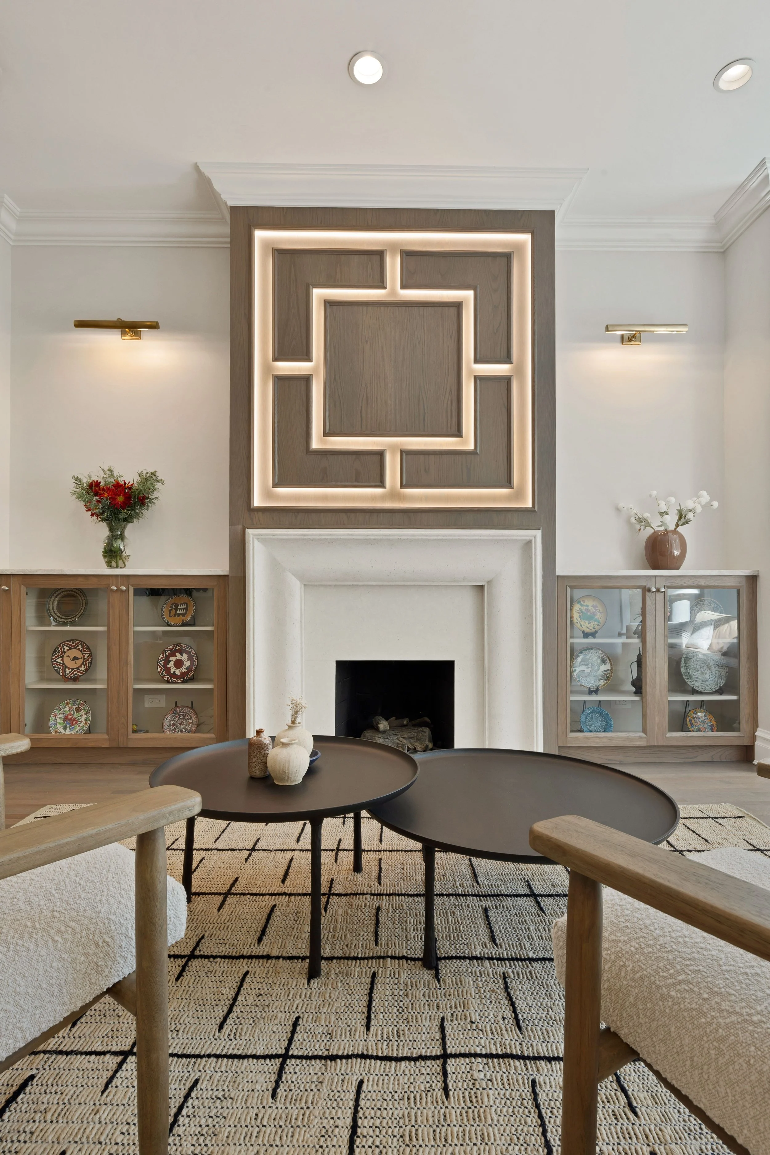 Living room with a white fireplace, decorative wall panel, two glass display cabinets, a round black coffee table, and wooden chairs on a beige rug.