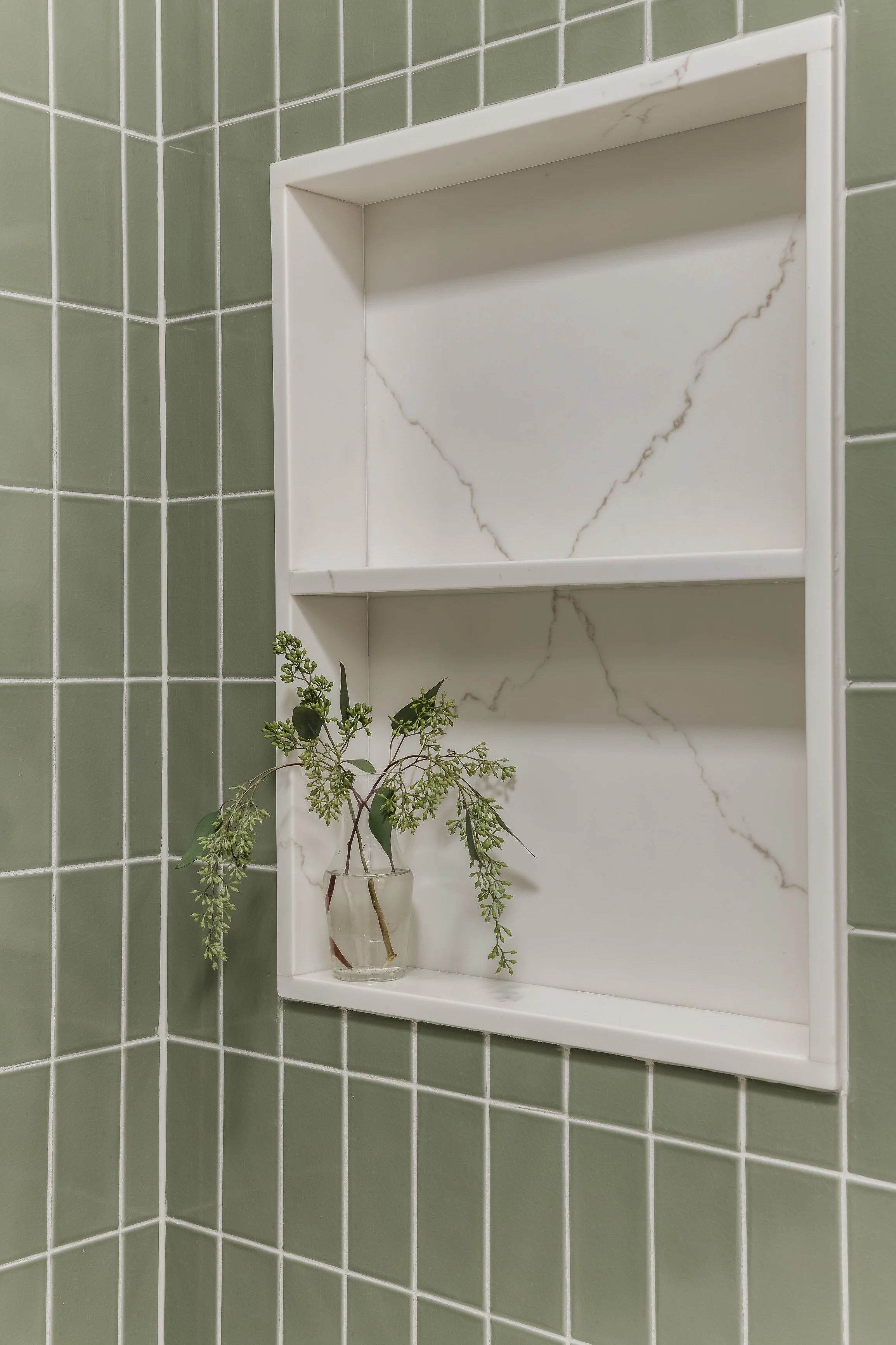 Green tiled bathroom wall with a white marble recessed shelf holding a small glass vase with green leafy sprigs.