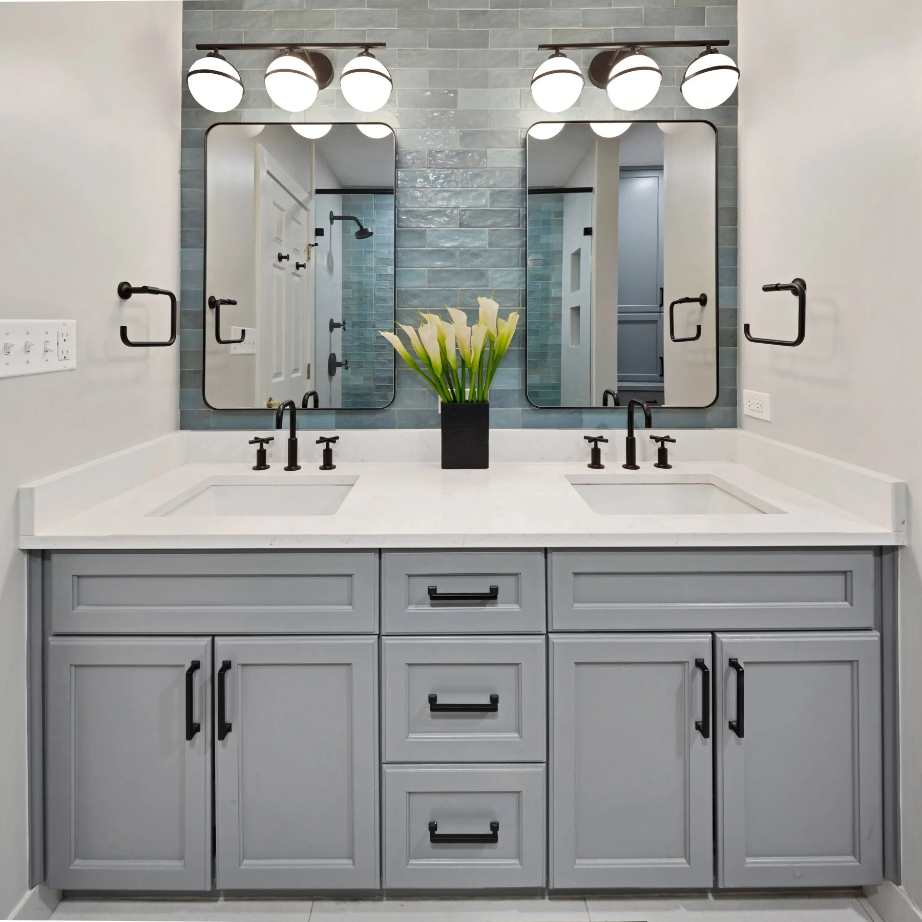Modern bathroom double vanity with light gray cabinets, a white countertop with two sinks, black fixtures, and large mirrors with black frames. There is a black vase with white flowers in the center and black towel bars on either side. The wall behin