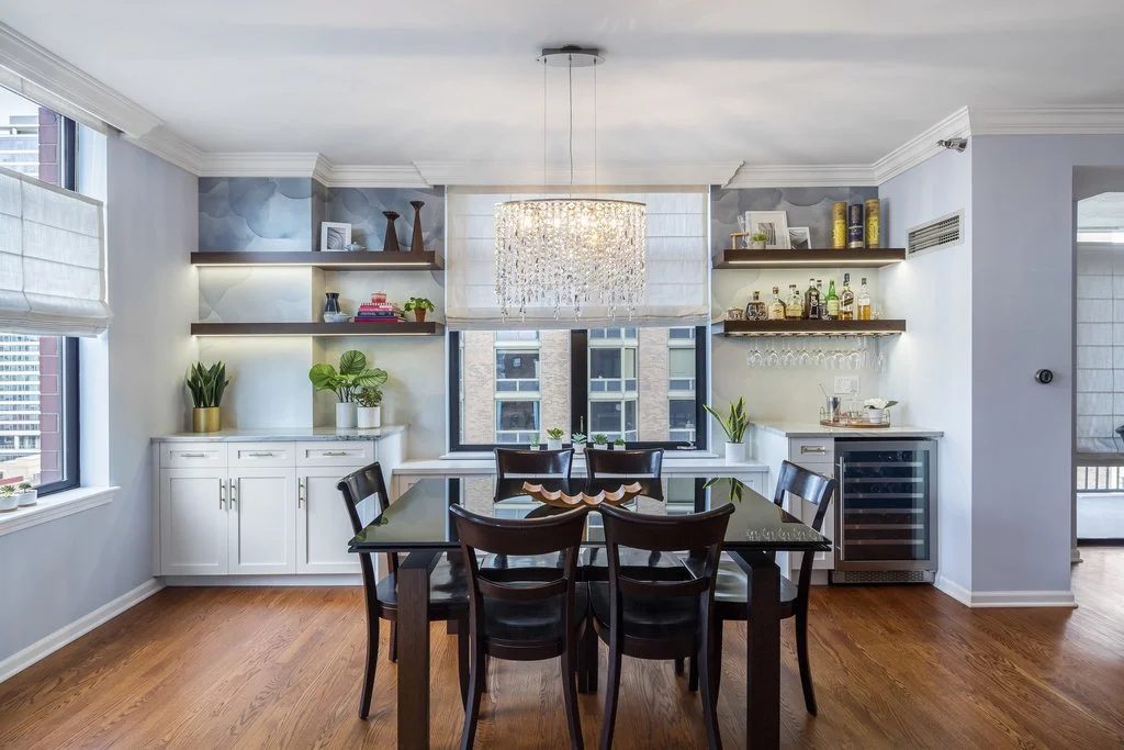 Modern dining room with a black table and six chairs, white cabinets, floating shelves with books and decorations, a chandelier, and windows with city views.