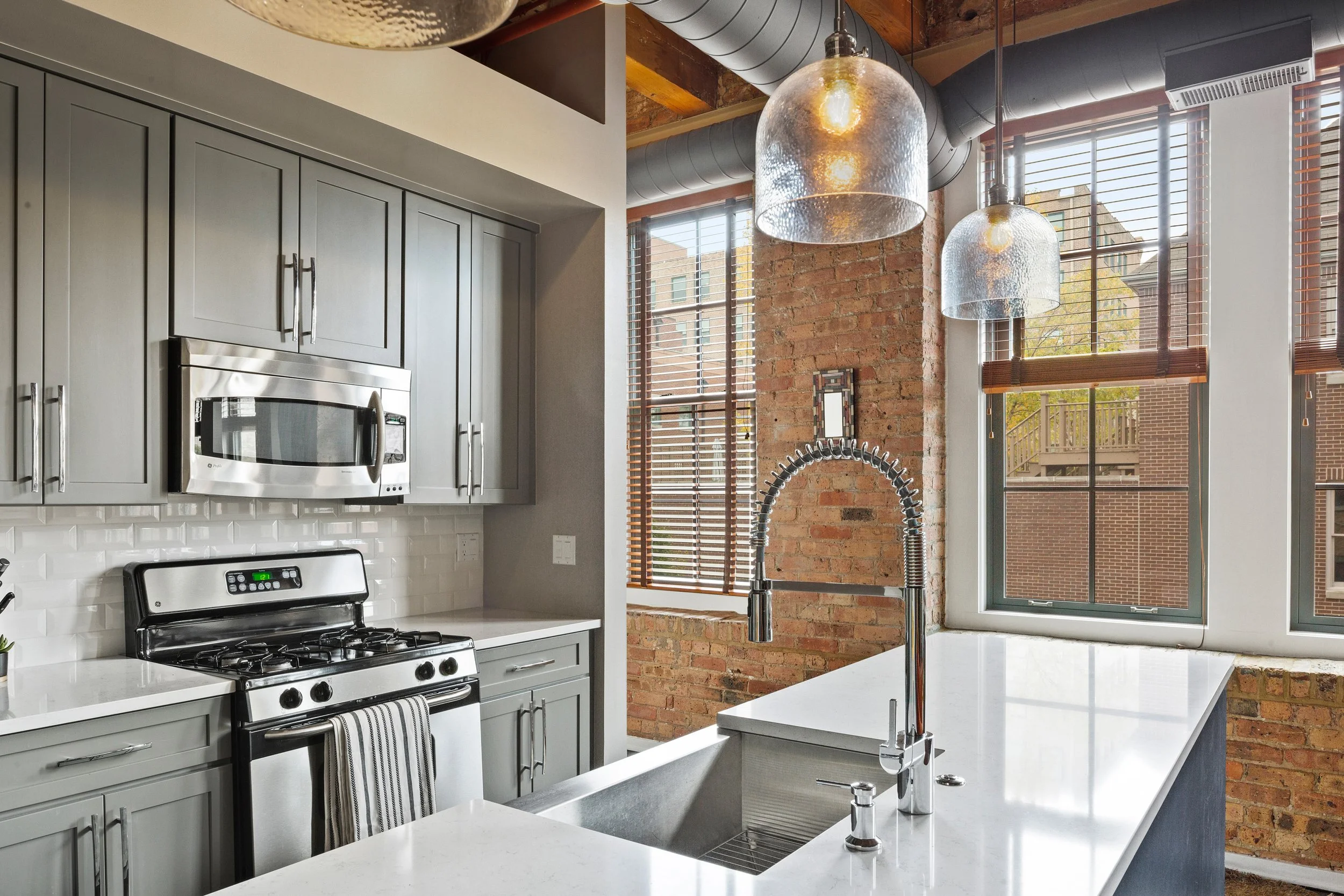Modern kitchen with gray cabinets, stainless steel microwave and stove, white countertops, and a brick accent wall with large windows and hanging pendant lights.