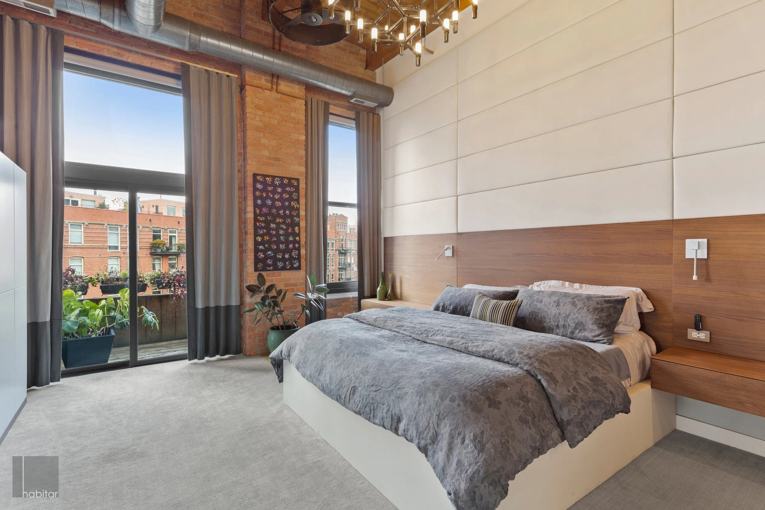 Modern bedroom with large bed, gray bedding, and decorative pillows. Wooden headboard and side table with green vases. Exposed brick wall with two windows, curtains, and a balcony with plants outside. Contemporary lighting fixture on ceiling.