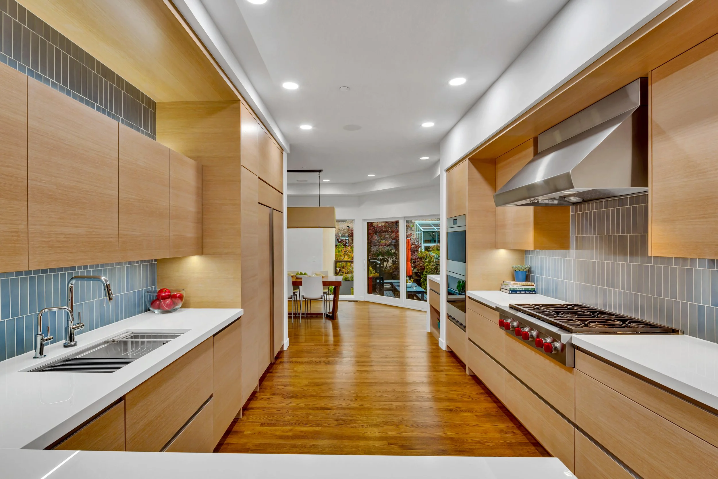 Modern kitchen with light wood cabinets, white countertops, blue tile backsplash, stainless steel oven and range hood, and hardwood flooring. Visible dining area with large windows and view of outdoor garden.