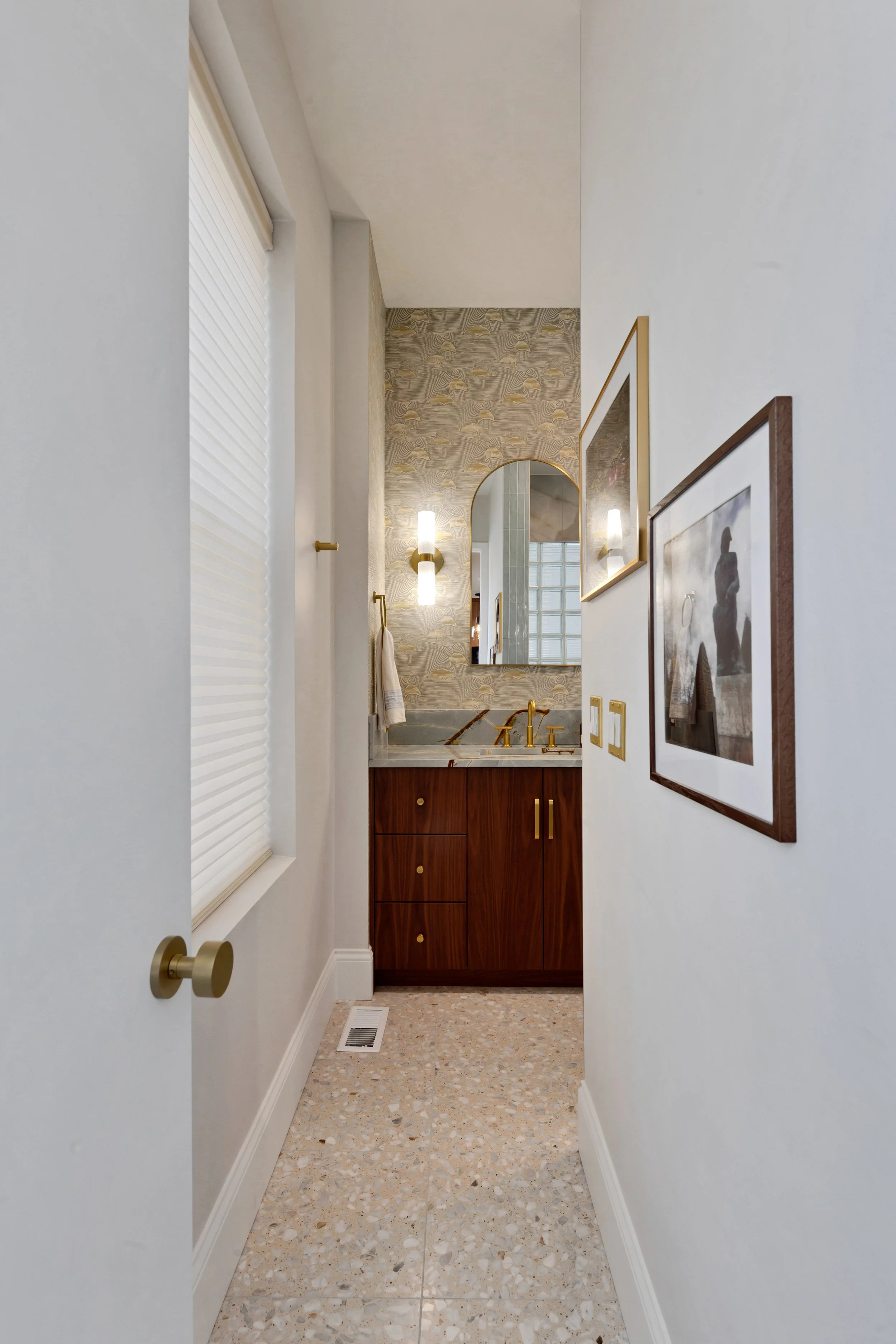 Bathroom with a vanity featuring a speckled marble countertop, gold fixtures, a round mirror, wall sconces, framed artwork, and patterned wallpaper.