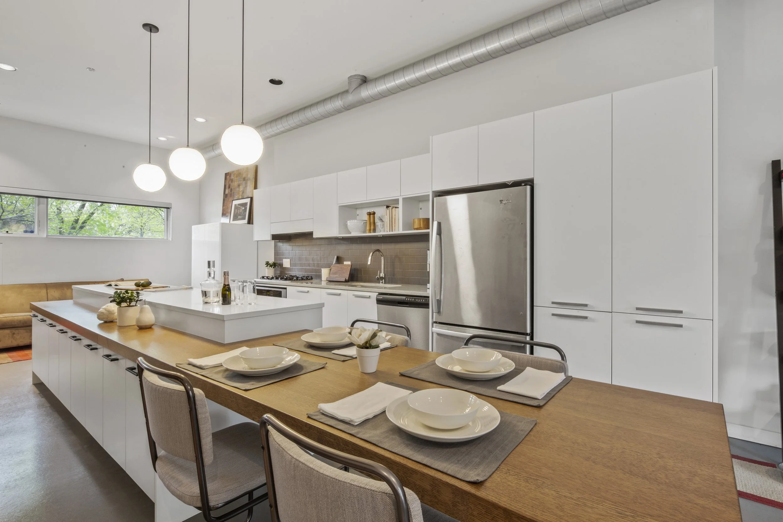 Modern kitchen with white cabinets, stainless steel refrigerator, and an island with a wooden countertop. The dining table set with four place settings is in the foreground, and a window with greenery outside is on the back wall.