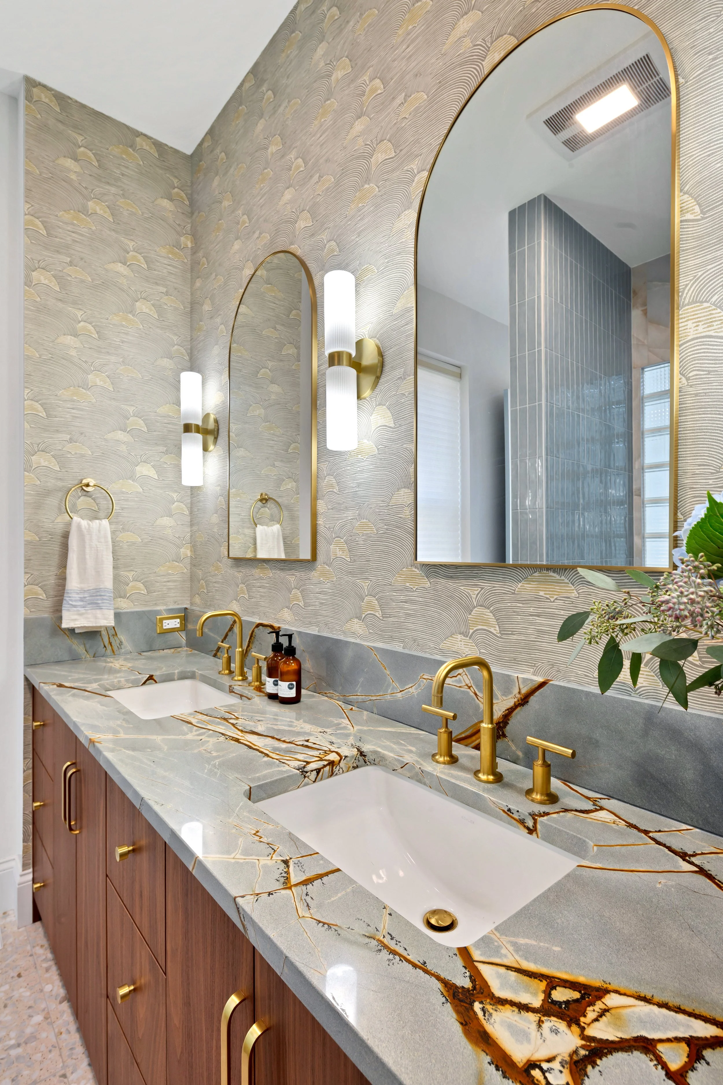 A modern bathroom double vanity with a marble countertop featuring gray, white, and gold veining, two white vessel sinks, and gold fixtures. There are two vertical mirrors with gold trim above each sink, flanked by wall sconces with gold bases and wh