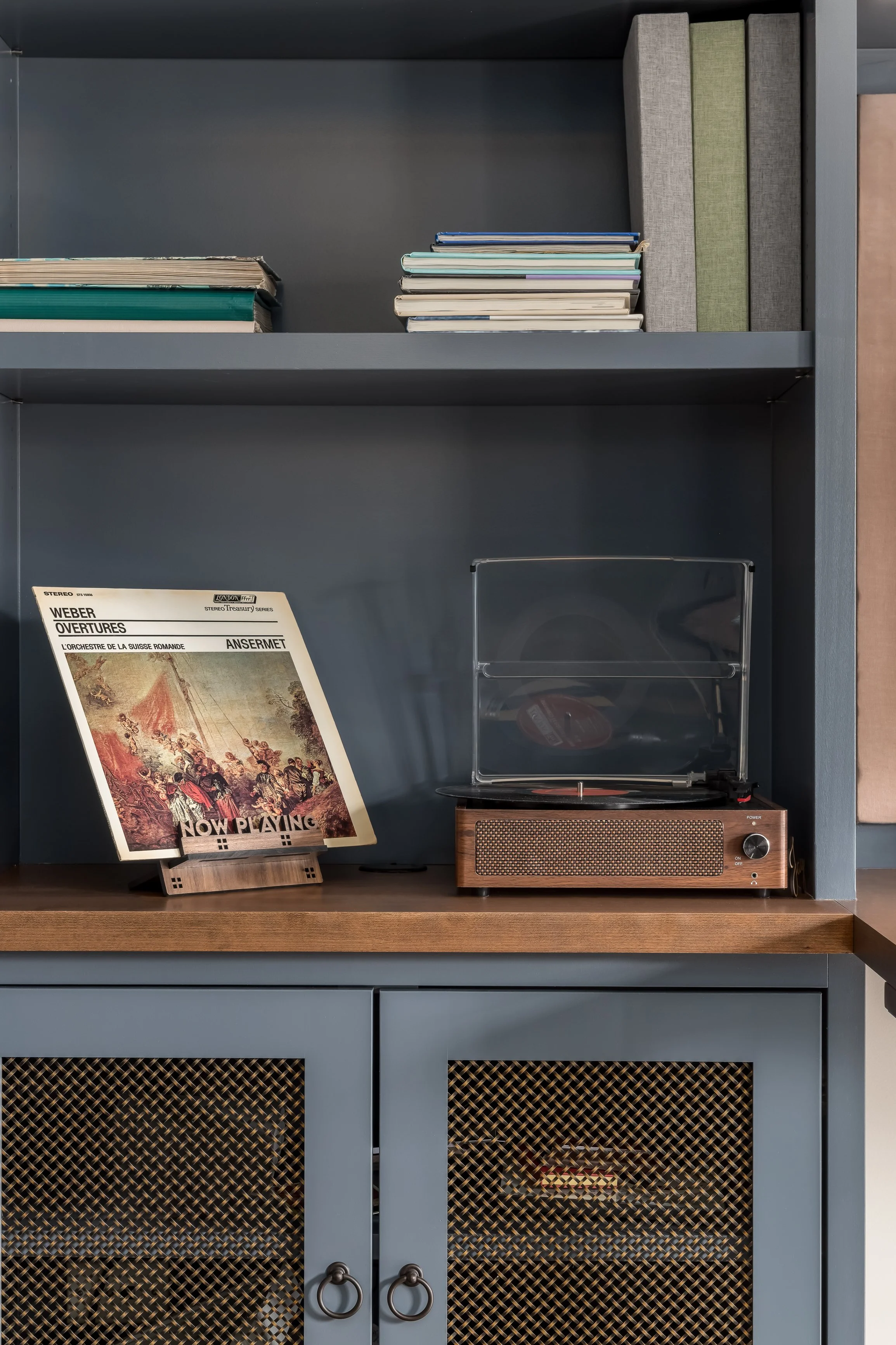 A vintage turntable with a clear dust cover, placed on a wooden cabinet, next to a vinyl record with a cover featuring a painting of a parade, on a blue shelving unit.