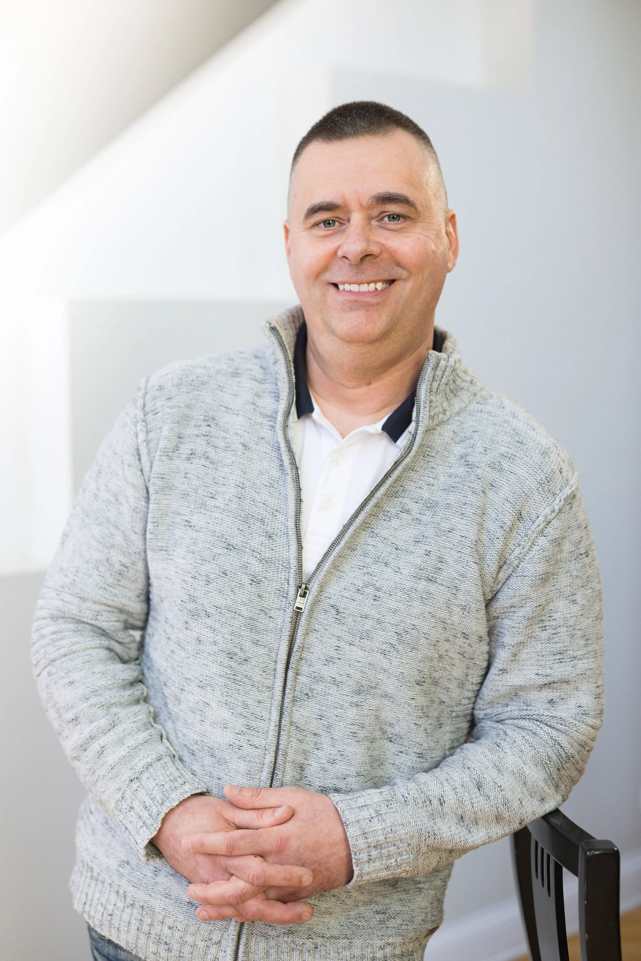 A smiling man wearing a light gray zip-up sweater and a white collared shirt posing indoors.
