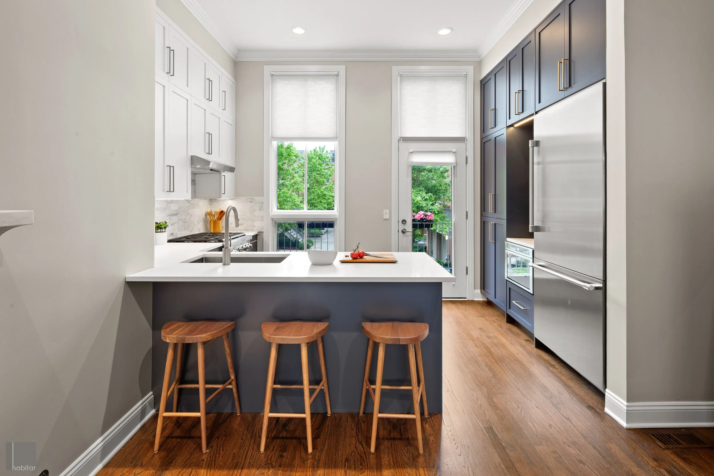 Modern kitchen with white upper cabinets, dark lower cabinets, stainless steel appliances, wooden bar stools, and hardwood flooring, with a view of trees outside through large windows.