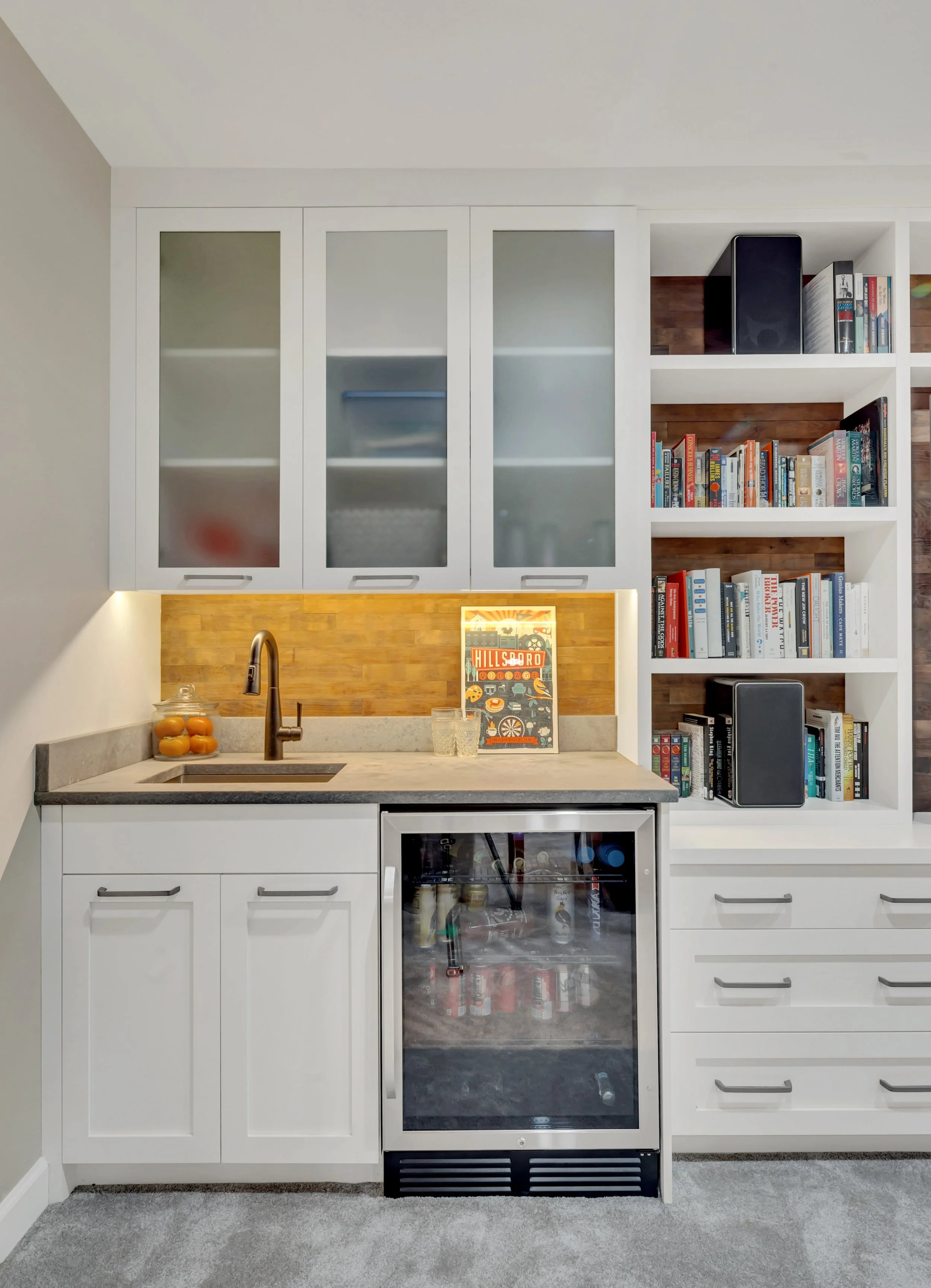 Small kitchen area with white cabinets, a wood backsplash, a small sink, a mini fridge with a glass door, open shelves with books and speakers, a wooden countertop, and a gray carpet.