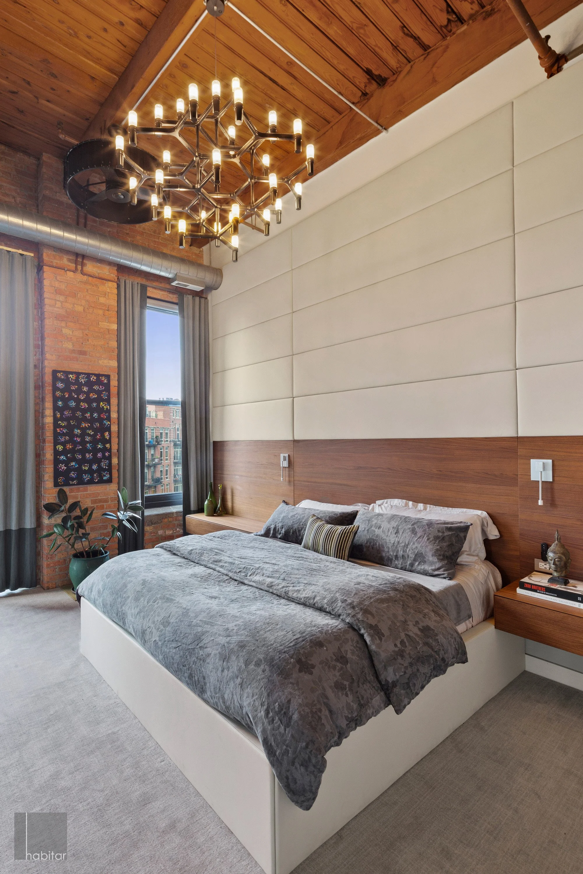 Modern bedroom with a large bed, gray bedding, wooden headboard, and a wall with padded panels. There is a black chandelier hanging from the wooden ceiling, a window with gray curtains, a potted plant, and a wall art piece.