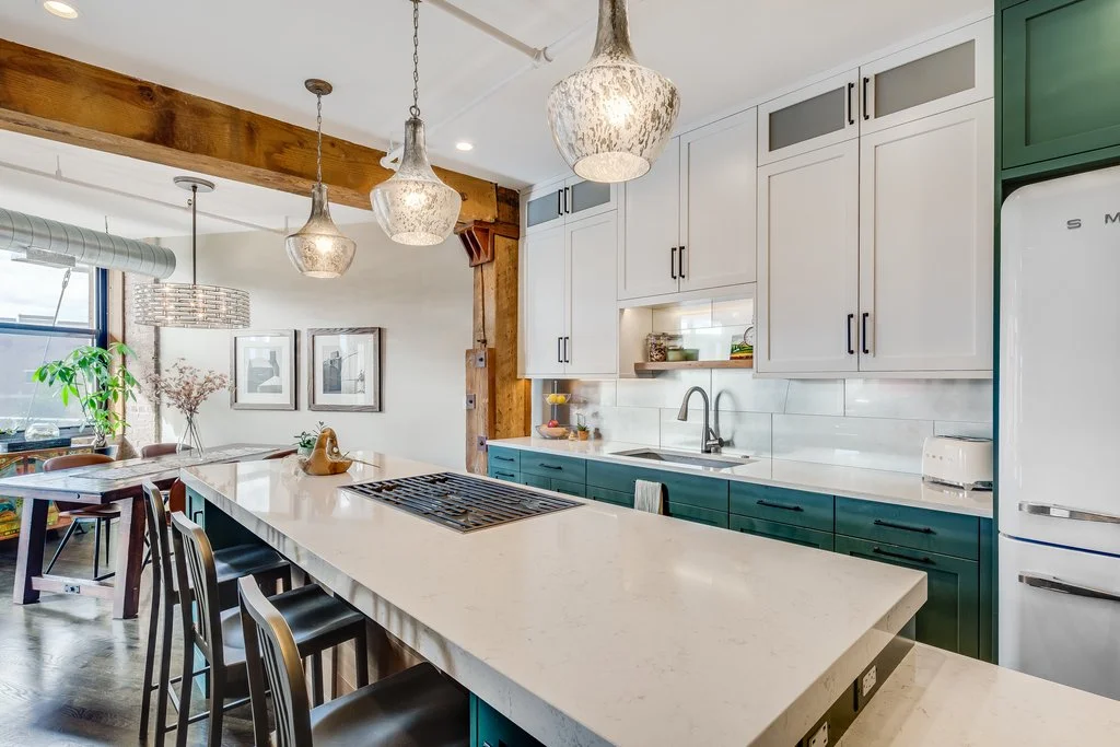 Modern kitchen with white and green cabinets, a large white island with four bar stools, hanging pendant lights, and a refrigerator, with a dining area nearby featuring a wooden table, chairs, and framed pictures on the wall.